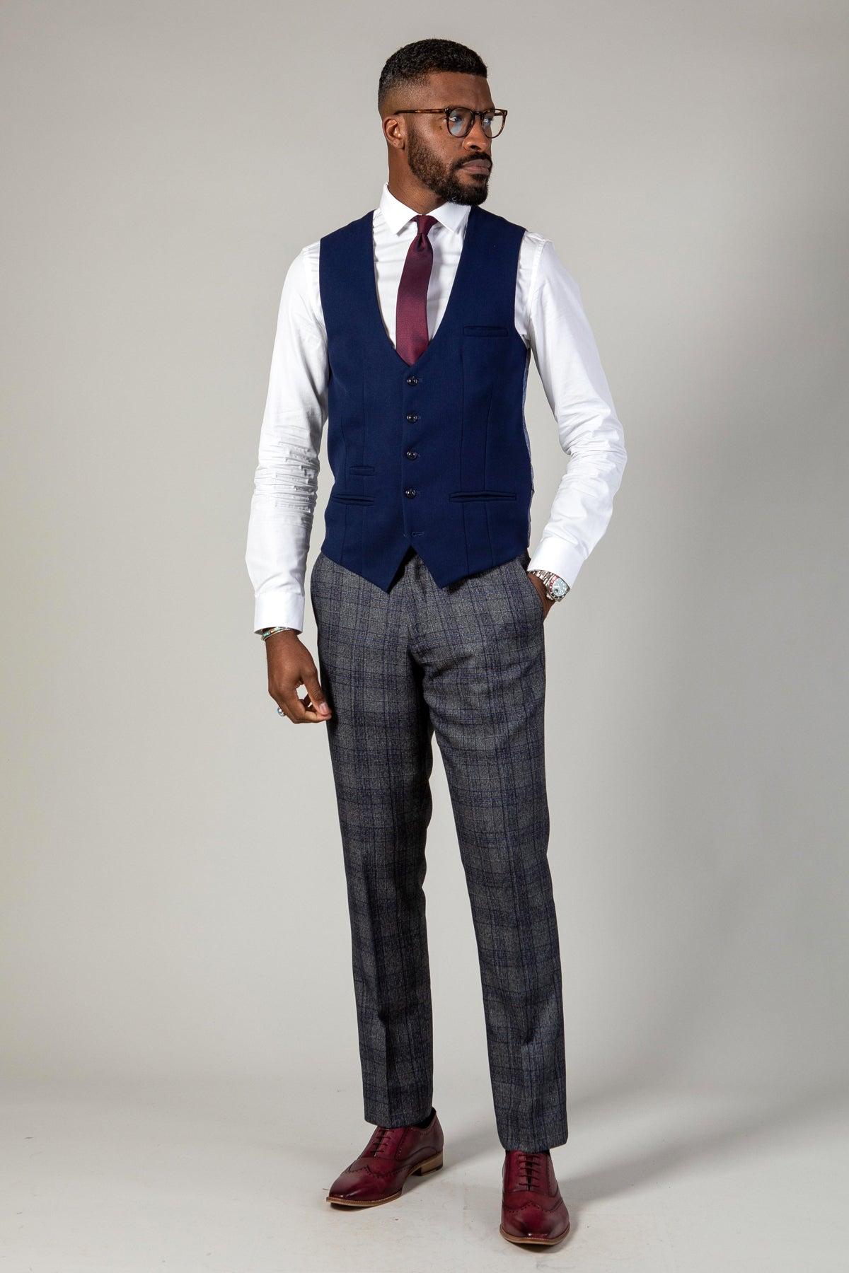 A man wearing the marcdarcy KELVIN Royal Blue Single Breasted Waistcoat with a white shirt, burgundy tie, and grey plaid pants, one hand in his pocket.