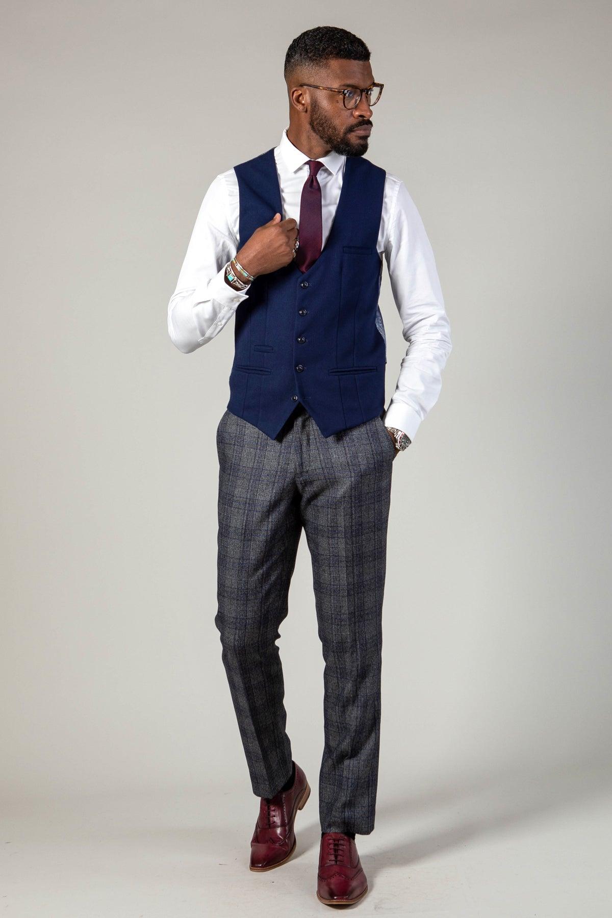 A man stands against a plain background, sporting glasses, a white shirt, dark red tie, marcdarcy's KELVIN Royal Blue Single Breasted Waistcoat, gray plaid trousers, burgundy shoes, a trimmed beard, and looking to his right.
