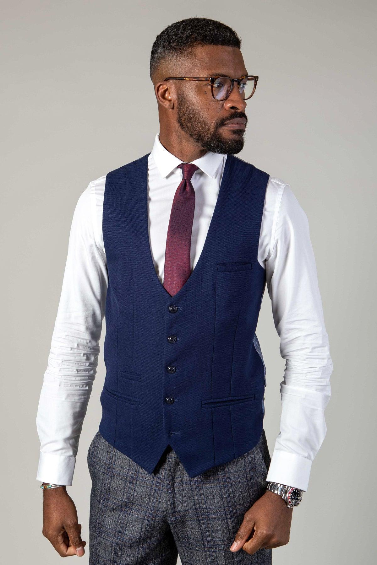 A Black man wearing glasses, a white dress shirt, a maroon tie, grey patterned pants, and the marcdarcy KELVIN Royal Blue Single Breasted Waistcoat stands against a light grey background.