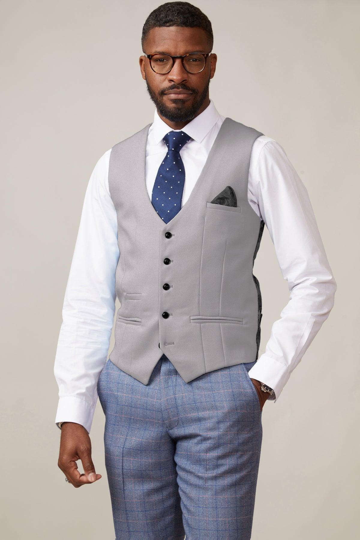 A bearded man with glasses wears the marcdarcy KELVIN Silver Single Breasted Waistcoat over a white shirt, navy polka dot tie, gray plaid trousers, and a watch, standing against a plain light background.