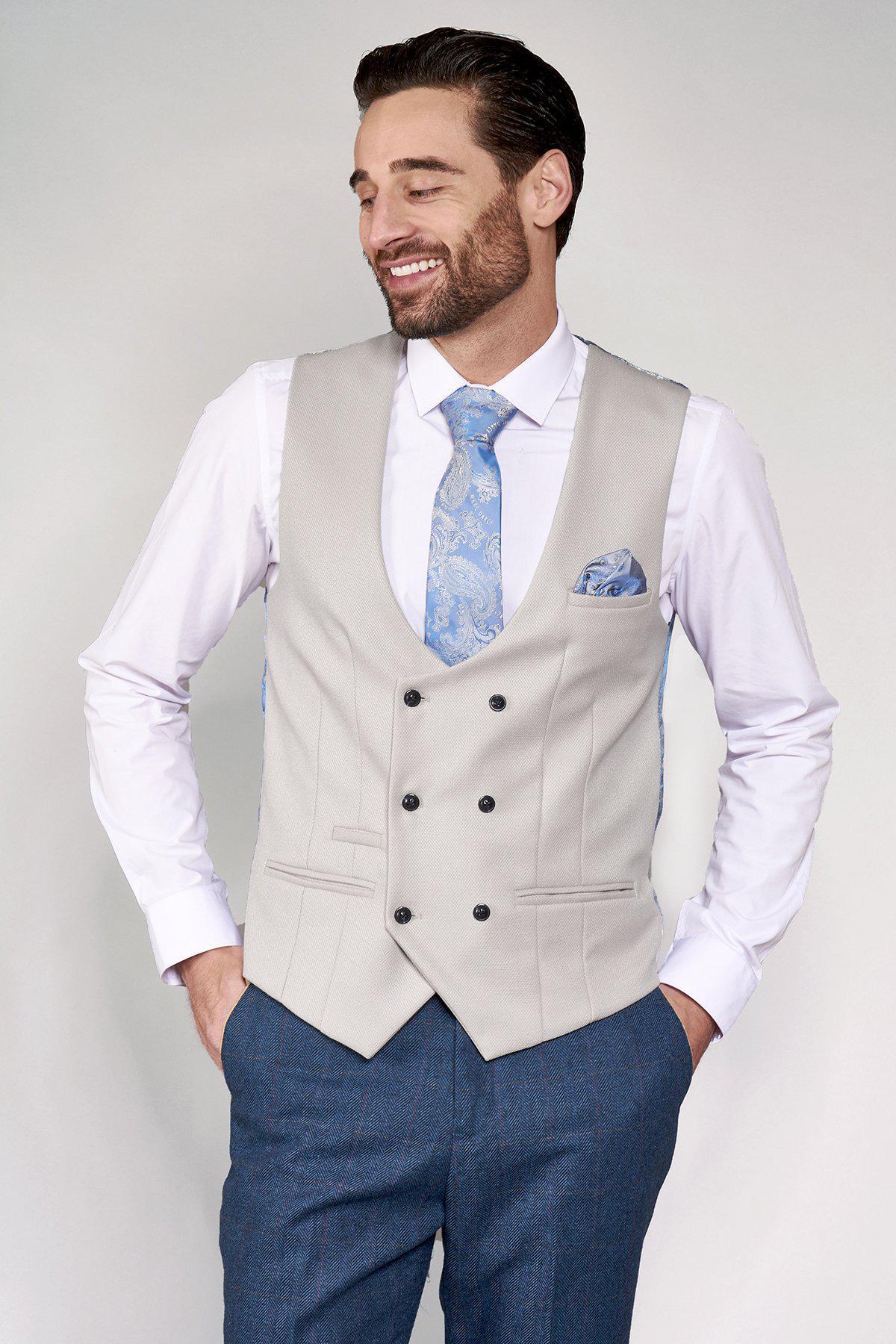 A man with dark hair and a beard smiles, looking left. He wears the marcdarcy DION Blue Tweed Check Suit with Kelvin Stone Waistcoat, white shirt, patterned blue tie, blue trousers, and matching pocket square.