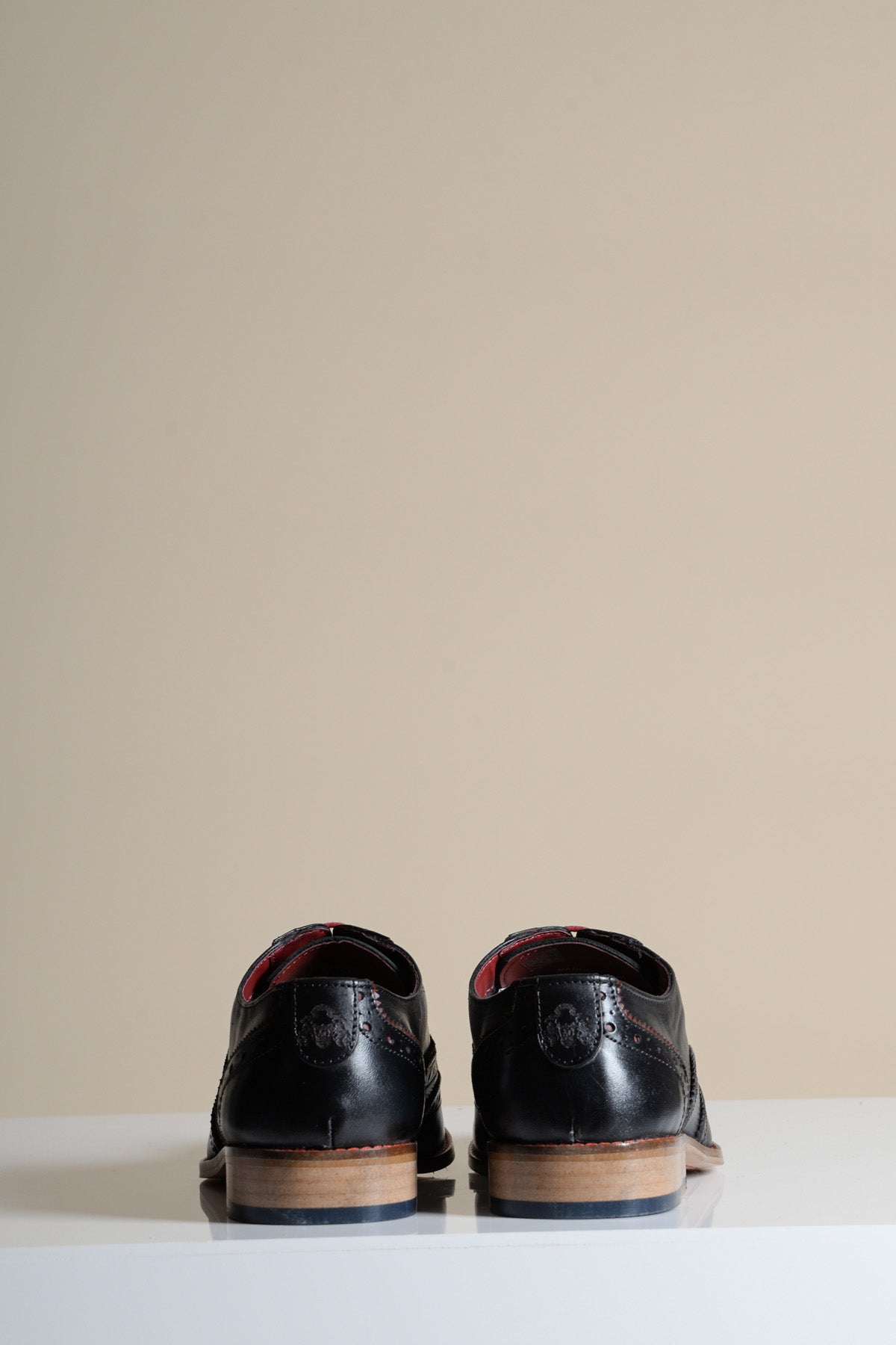 The MARCO - Black Bordeaux Leather Wingtip Oxford Shoe by Marc Darcy, featuring full grain leather and brown soles, is shown facing backward on a white surface with a beige background.