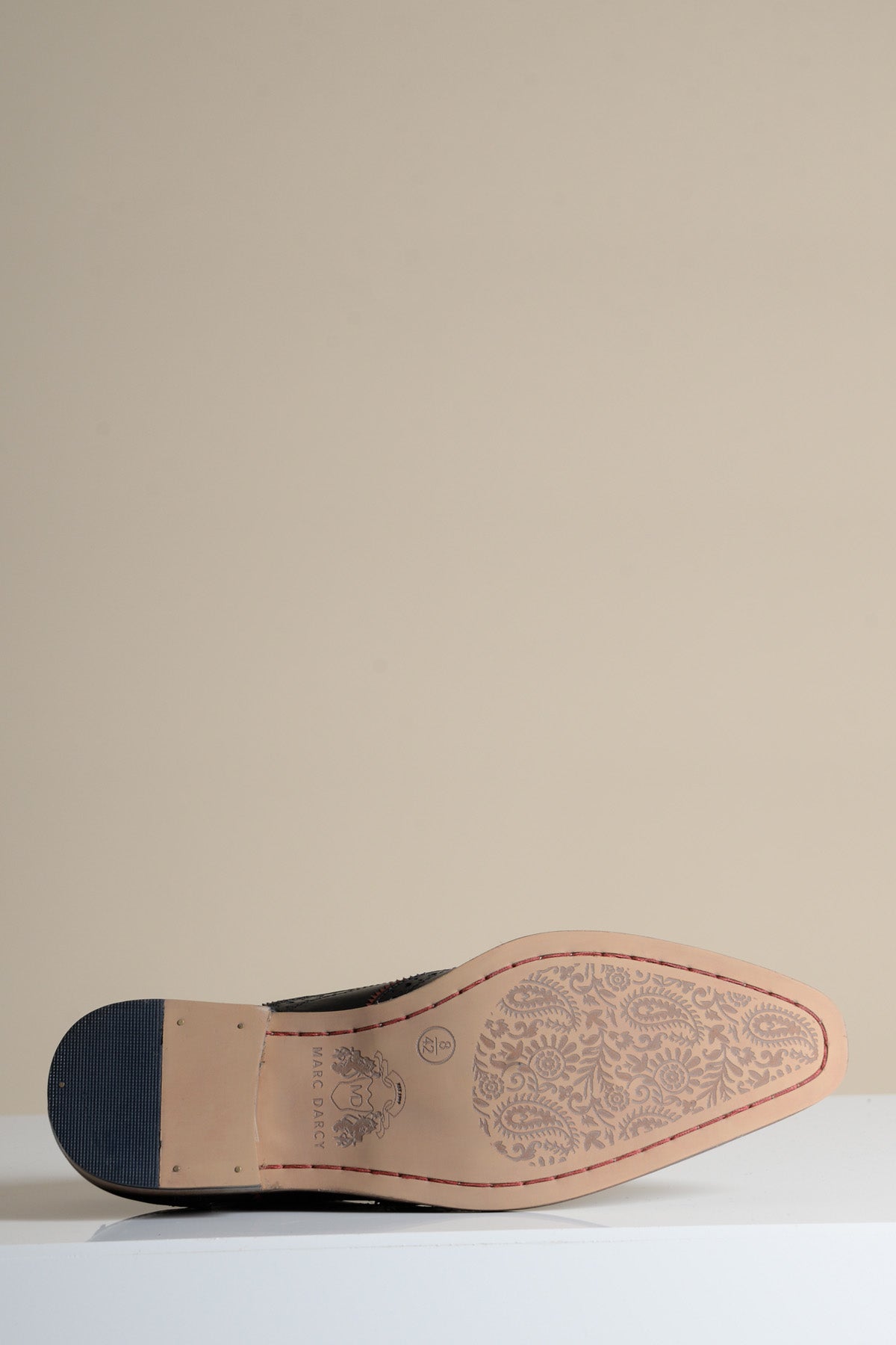A single MARCO Black Bordeaux Leather Wingtip Oxford Shoe by Marc Darcy rests on its side, highlighting the sole’s detailed floral pattern etched into the light brown full grain leather against a plain beige background.