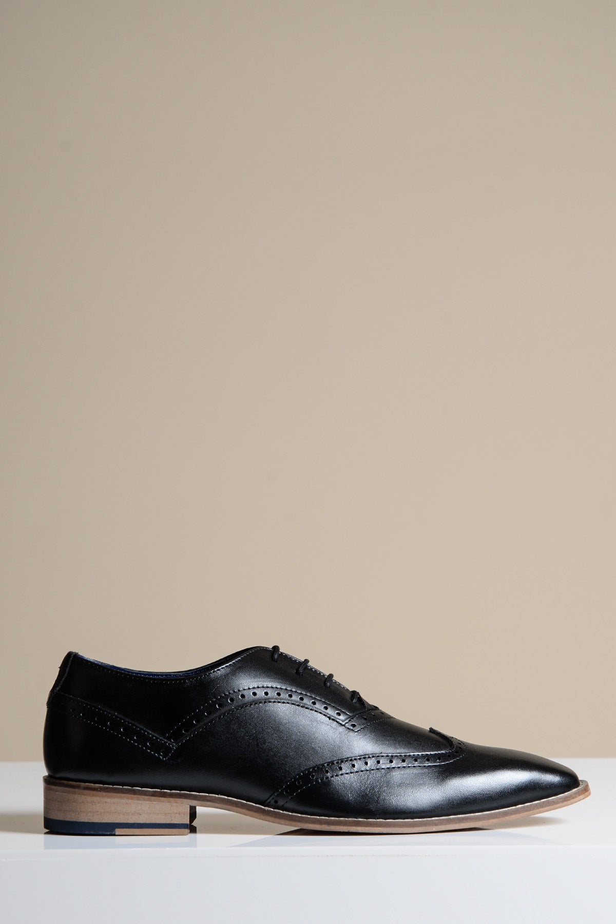 The Marc Darcy DAWSON is a black wingtip oxford brogue shoe with decorative stitching, laces, and a low wooden heel, crafted from full grain leather and shown on a white surface against a beige background.