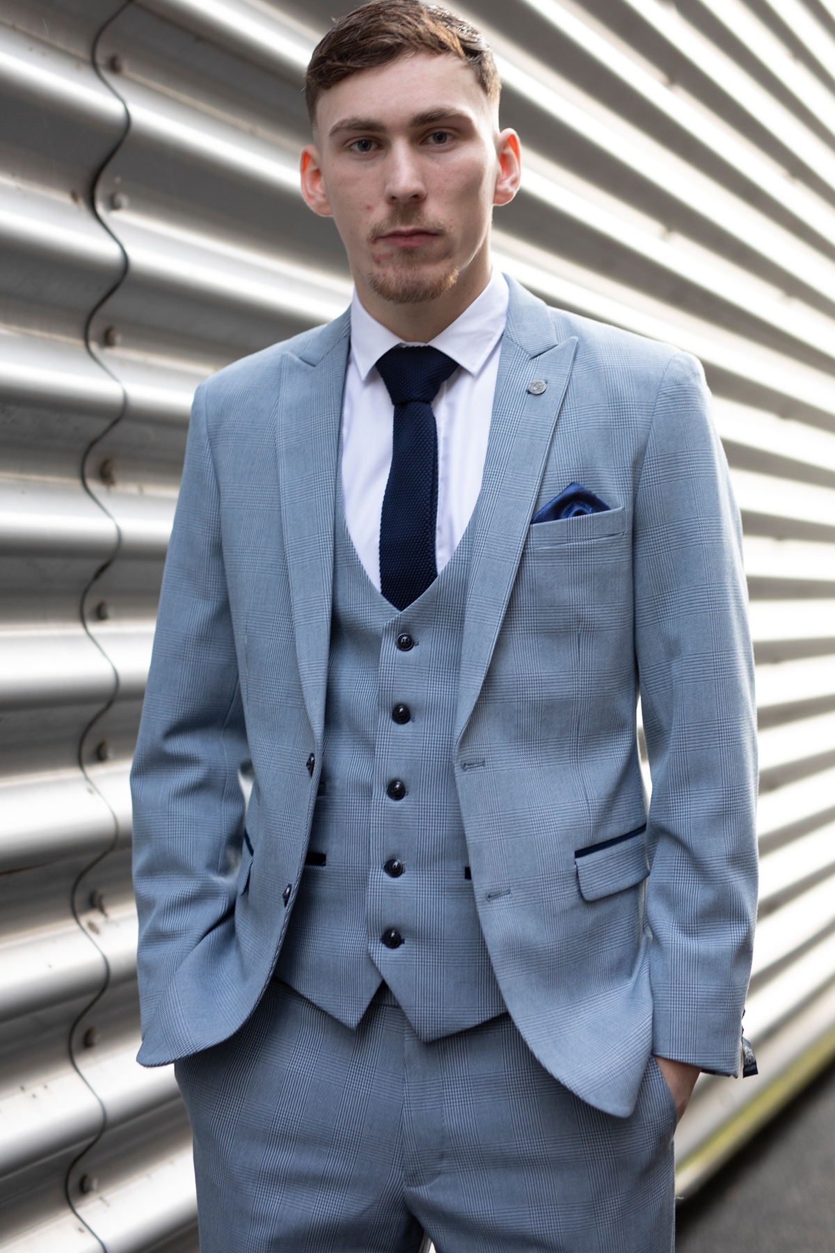 A young man models Marc Darcy’s The Everton Collection | James Garner In The BROMLEY Sky Blue Suit, standing hands in pockets before a corrugated metal wall and looking directly at the camera.