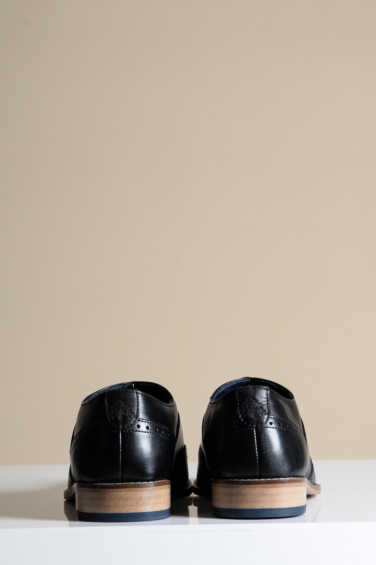 A pair of Marc Darcy DAWSON Black Wingtip Oxford Brogue Shoes, made from full grain leather, shown from the back on a white surface with a beige background.