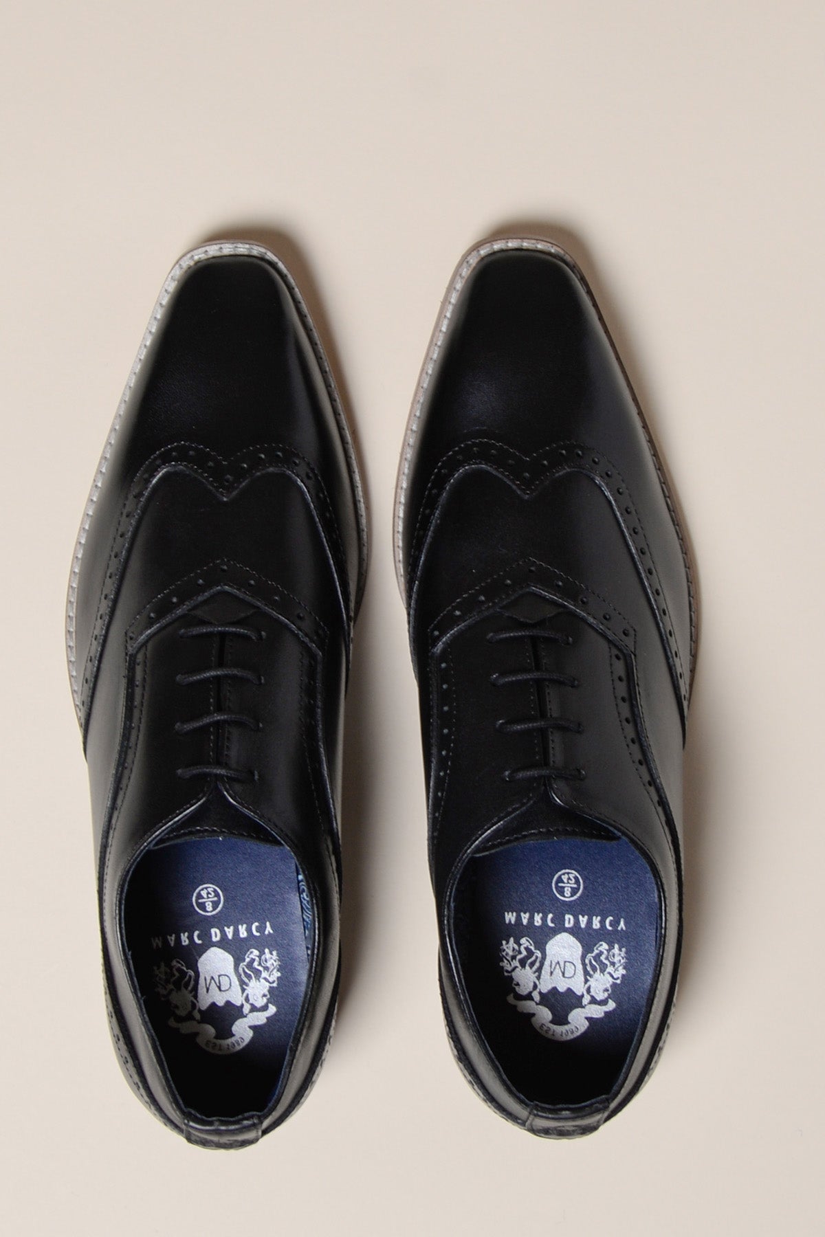 A pair of Marc Darcy DAWSON Black Wingtip Oxford Brogue Shoes, featuring decorative stitching, laces, and pointed toes, shown from above on a light beige background with insoles displaying a crest and design.