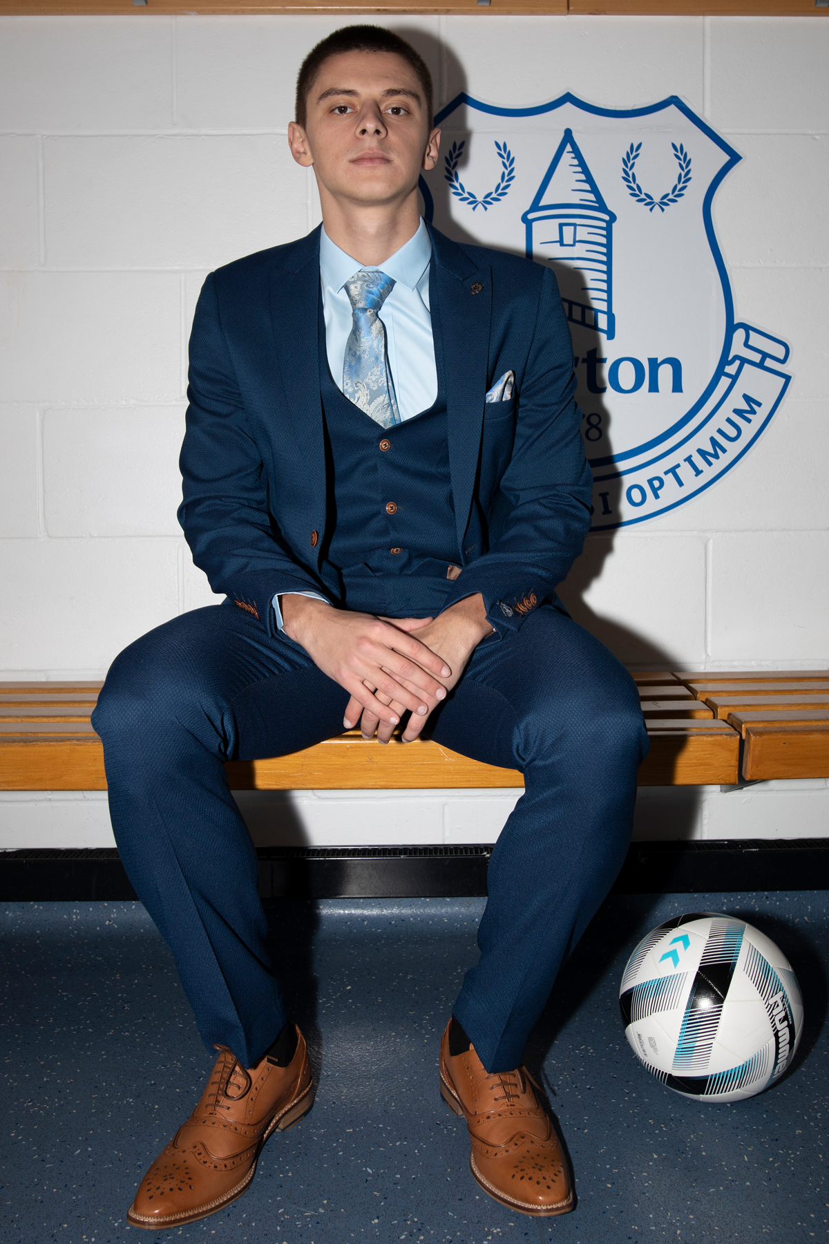 Vitalii Mykolenko models The Everton Collection | MAX Royal Blue Suit by marcdarcy, seated confidently on a wooden bench with hands clasped. A soccer ball sits nearby, and a blue and white Everton club emblem is on the wall behind him.