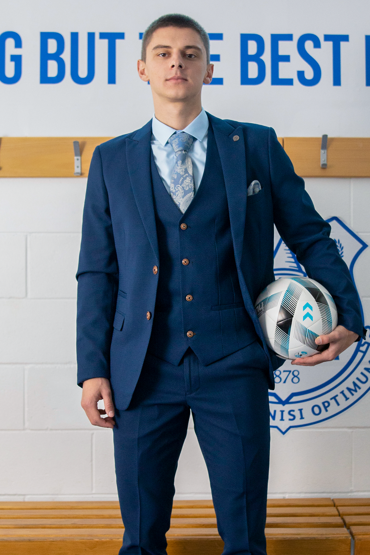 A young man models The Everton Collection | Vitalii Mykolenko In The MAX Royal Blue Suit by marcdarcy, standing indoors with a soccer ball under one arm, against a wall with hooks and a partially visible sign and emblem.