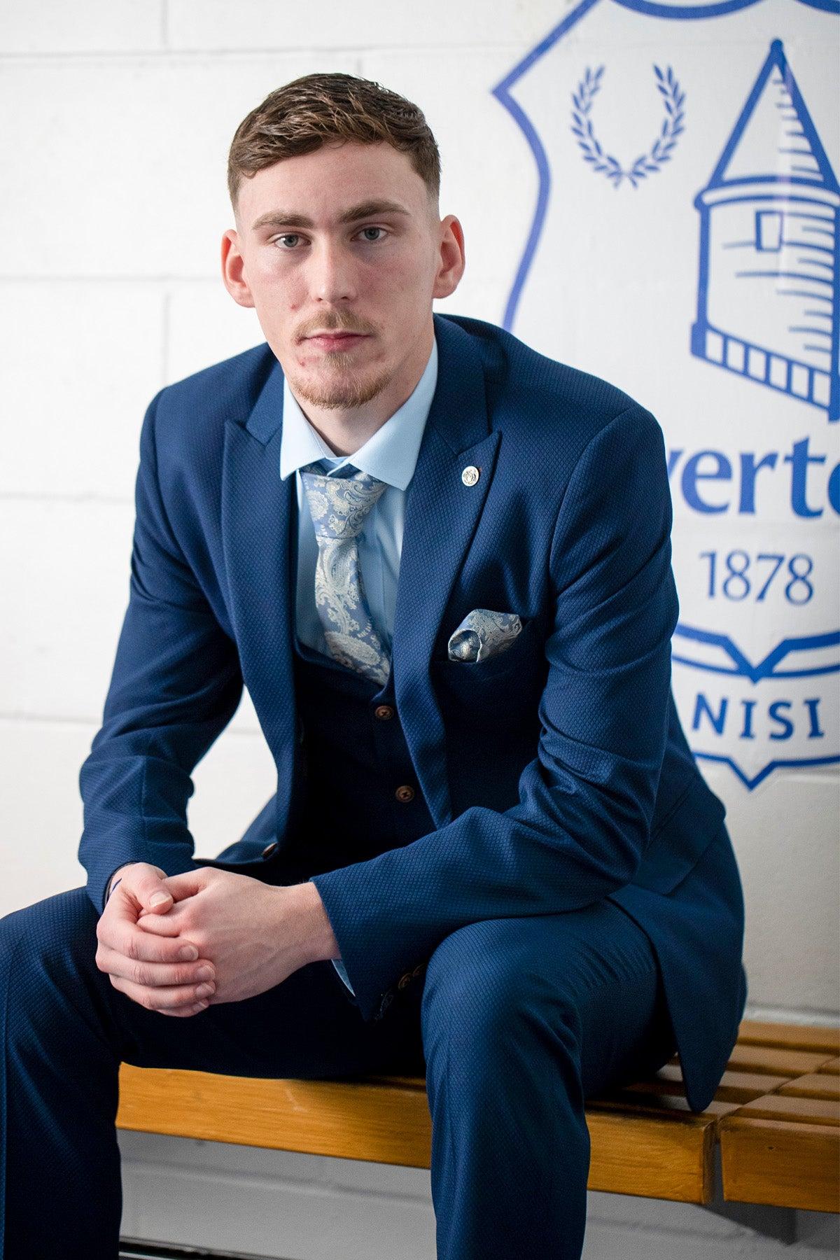 A young man wears the marcdarcy The Everton Collection | James Garner In The MAX Royal Blue Suit, sitting on a wooden bench before a white wall with part of the blue Everton football club logo behind him.