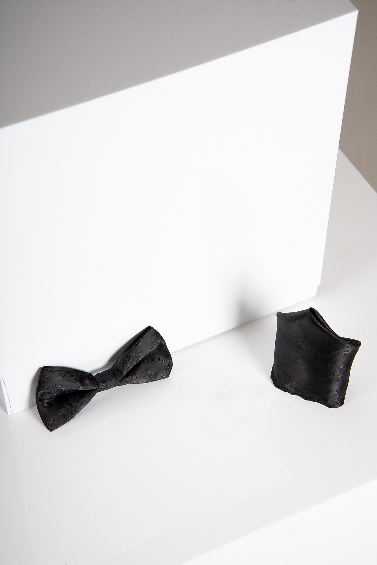 The Accessory MD PAISLEY Black Paisley Bow Tie and Pocket Square Set is displayed on a glossy white surface beside a white rectangular block.