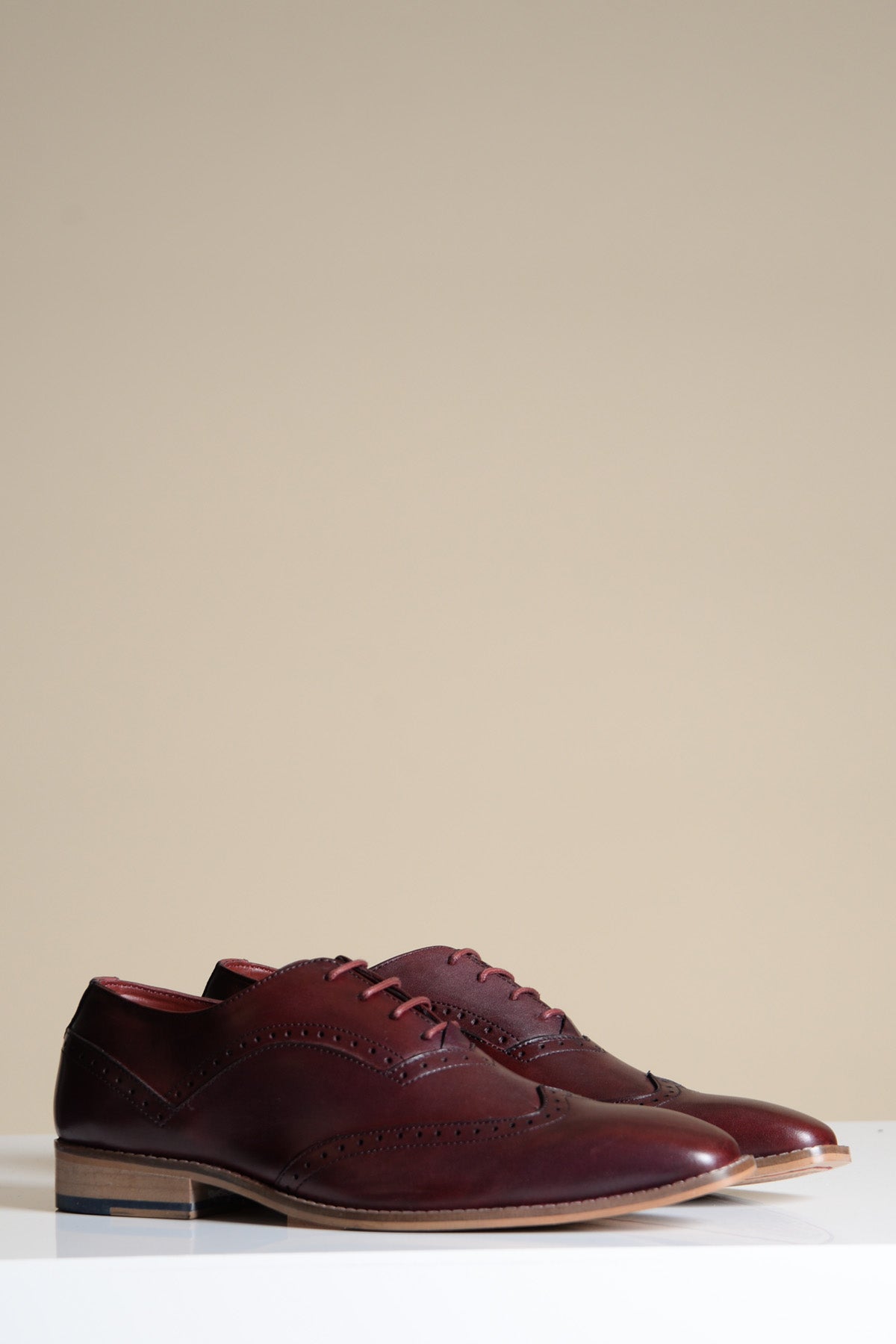 A pair of Marc Darcy DAWSON Bordeaux Burgundy Wingtip Oxford Brogue Shoes, featuring laces and decorative stitching, are displayed on a white surface with a beige background.