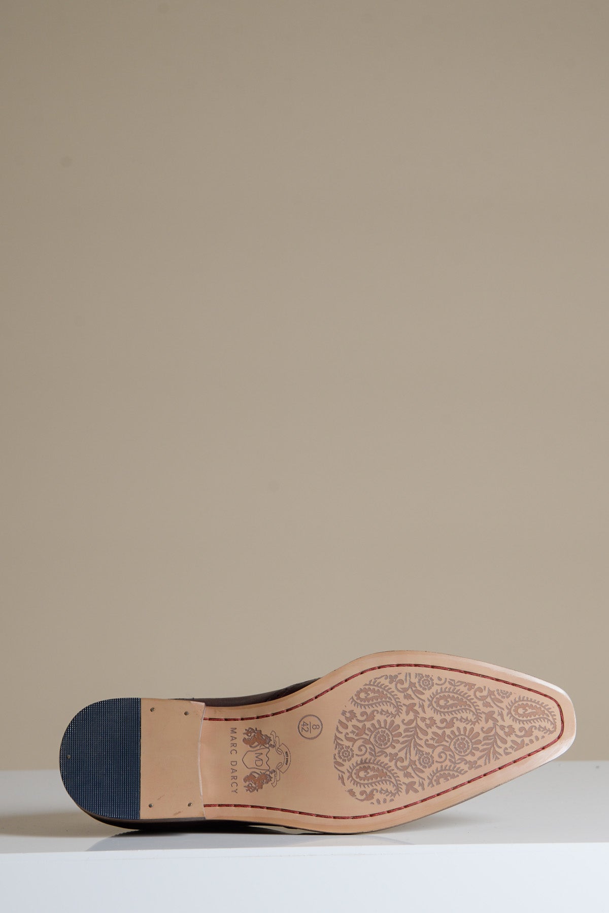A close-up of the sole of the Marc Darcy DAWSON Bordeaux Burgundy Wingtip Oxford Brogue shows a blue heel and intricate floral patterns etched into light brown full grain leather, set against a plain beige background.
