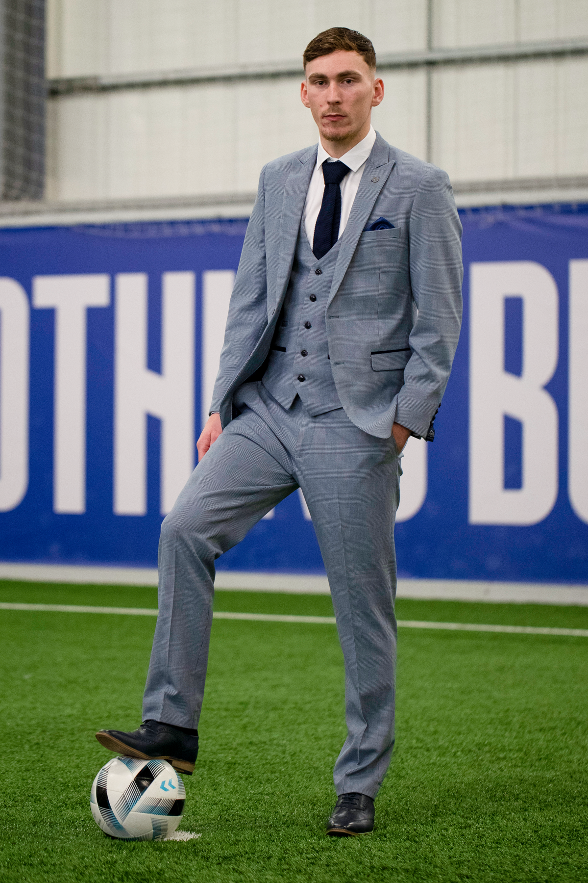 A man in the Marc Darcy Everton Collection "James Garner In The BROMLEY Sky Blue Suit" stands on indoor artificial turf, confidently resting one foot on a soccer ball, with a large blue banner in the background.