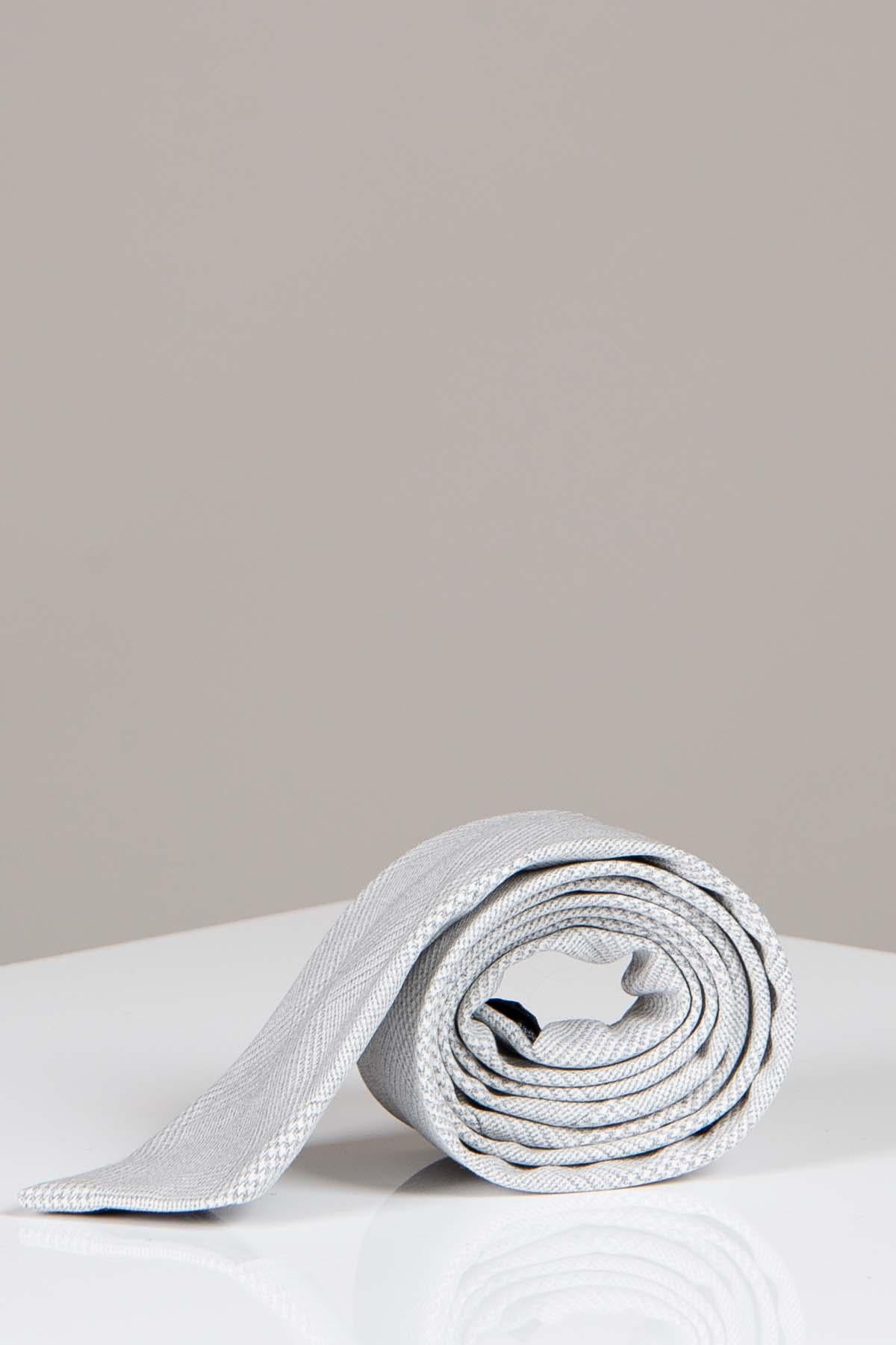 The BROMLEY - Stone Check Tie by Accessory, featuring a light gray check pattern, is rolled up on a white surface against a neutral background.