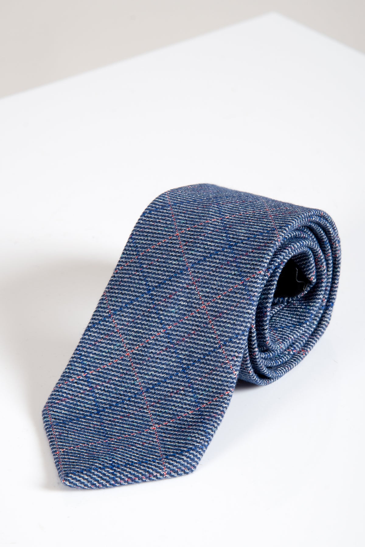 The HILTON Blue Check Tweed Tie by Accessory is a men’s dark blue necktie featuring a subtle plaid of thin red and white lines, shown rolled up on a white background with a soft shadow.