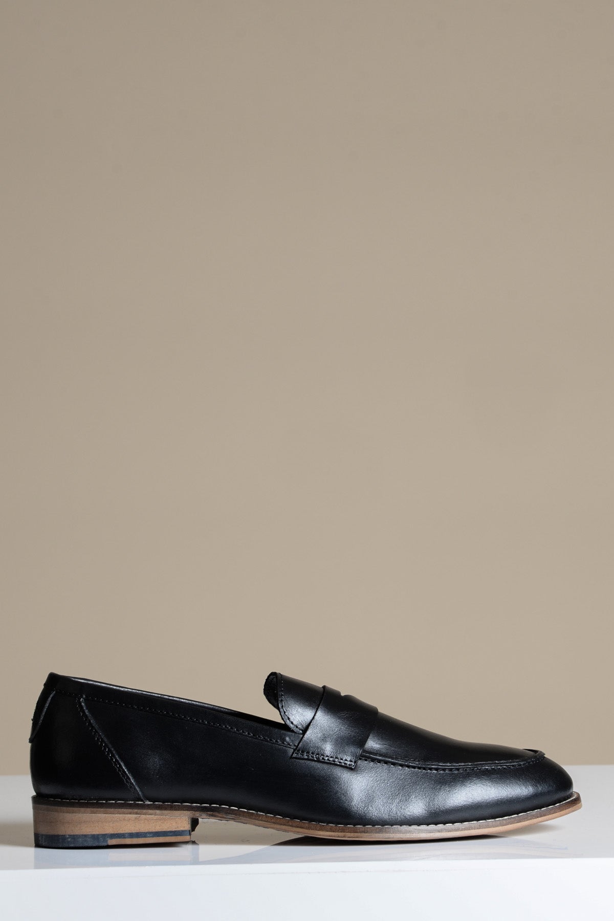 The Marc Darcy JASPER Black Leather Penny Loafer Shoe, featuring a low wooden heel and hand-stitched details, is showcased in side profile on a white surface with a beige background.