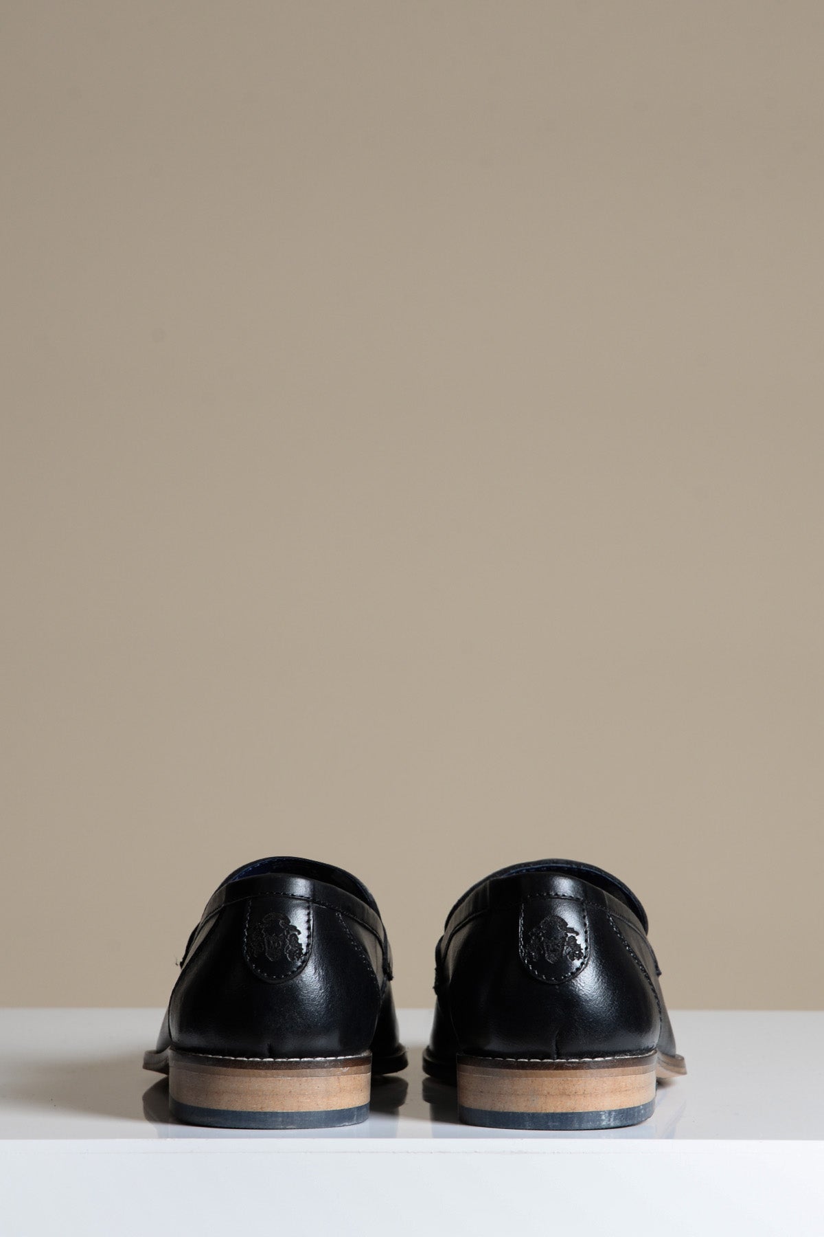 The JASPER Black Leather Penny Loafer Shoes by Marc Darcy are shown with the heels facing forward on a white surface, set against a beige background.