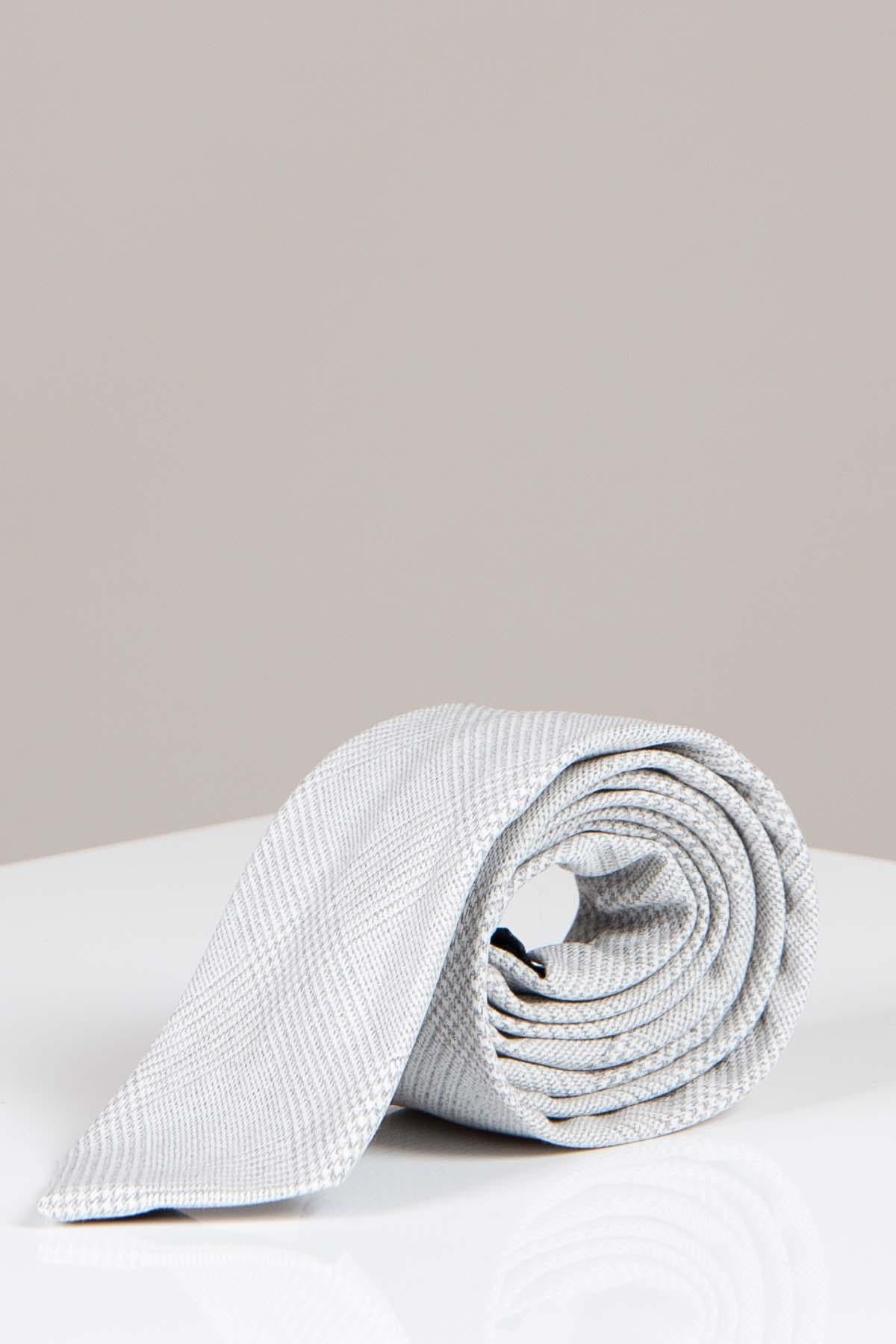 The Accessory BROMLEY - Stone Check Tie, featuring a light gray Prince of Wales pattern, is neatly rolled on a glossy white surface, making it an ideal formalwear choice against a plain light beige background.