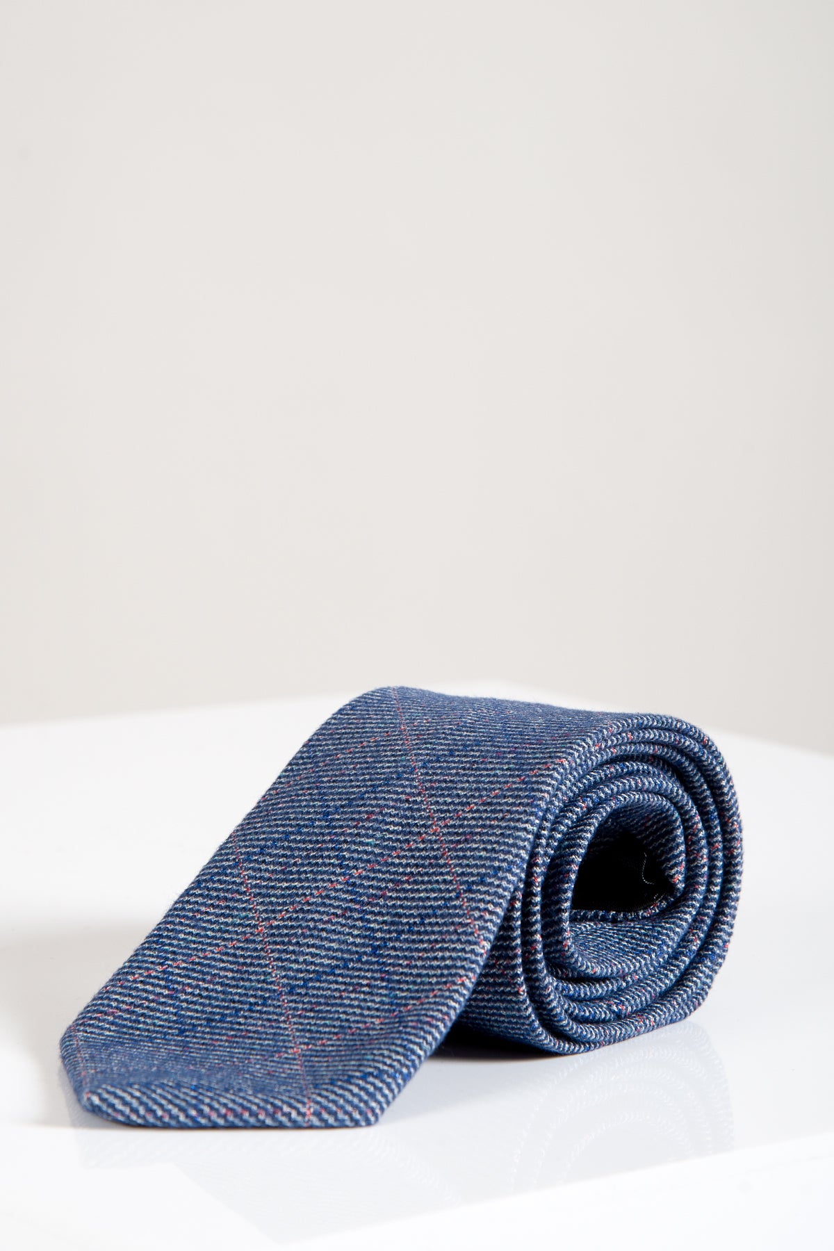 The HILTON - Blue Check Tweed Tie by Accessory, featuring a blue check pattern with subtle red lines, is neatly rolled on a white surface against a plain light background.