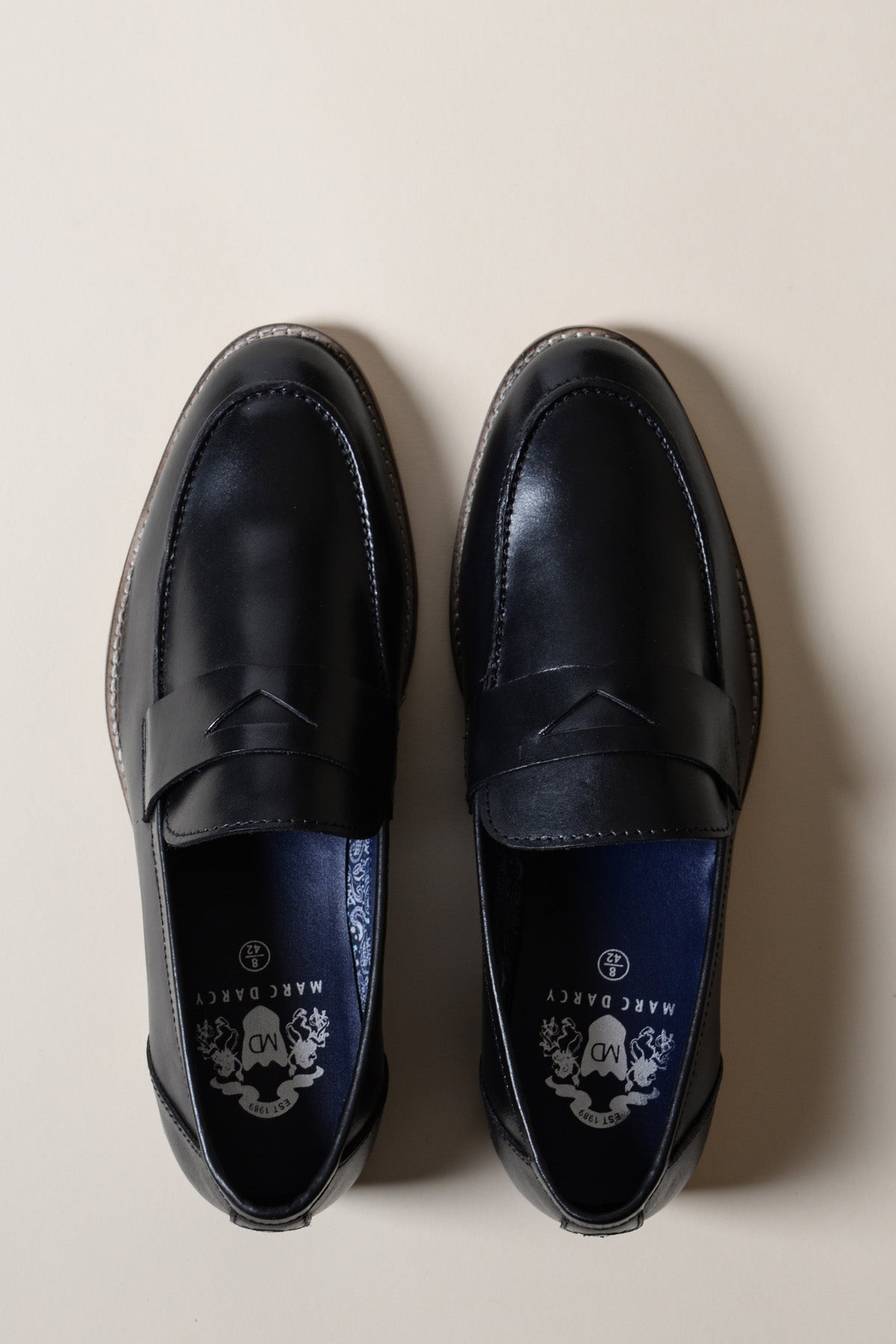 The JASPER Black Leather Penny Loafer Shoe by Marc Darcy is displayed side by side on a beige background, featuring a penny slot detail and dark blue insoles adorned with a decorative white logo and crest.