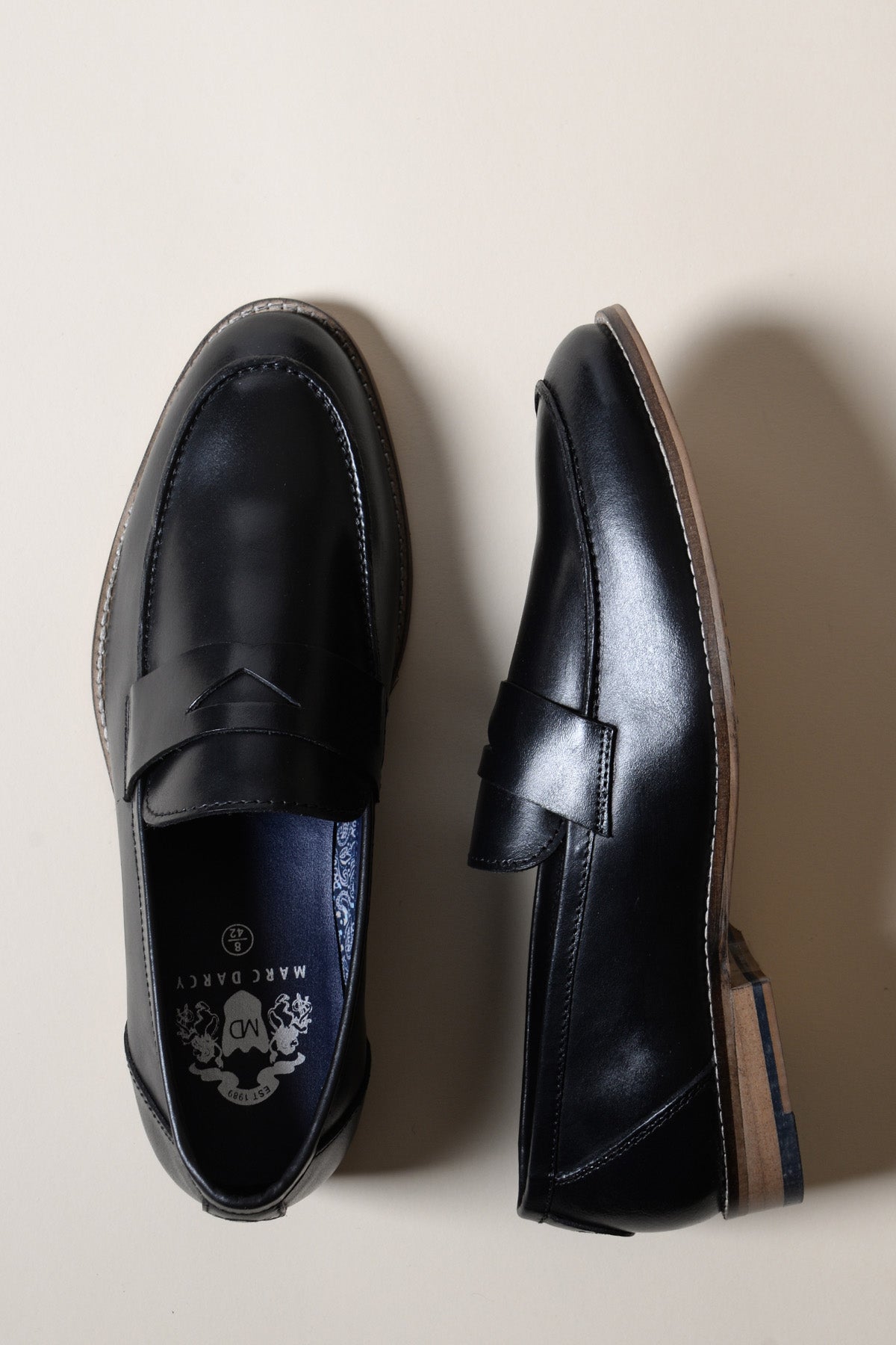 The Marc Darcy JASPER Black Leather Penny Loafer Shoes, with brown soles and fine stitching, are shown on a light beige background—one top view and one side view highlight their classic profile and craftsmanship.
