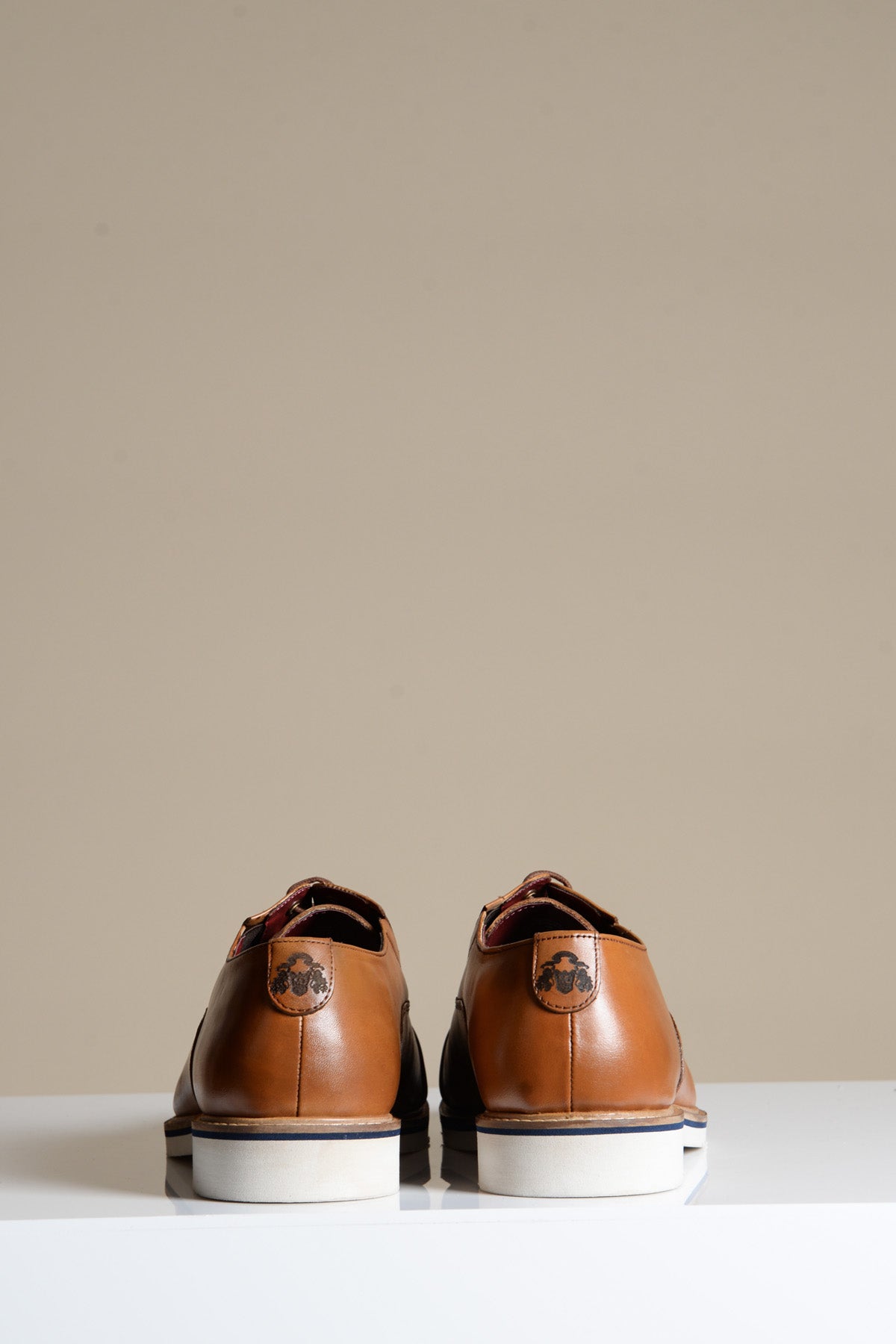 The GAVIN Tan Leather Cap Toe Oxford Shoes by Marc Darcy are shown from the back on a beige background, each featuring a small circular logo near the heel and white soles for a refined, modern look.