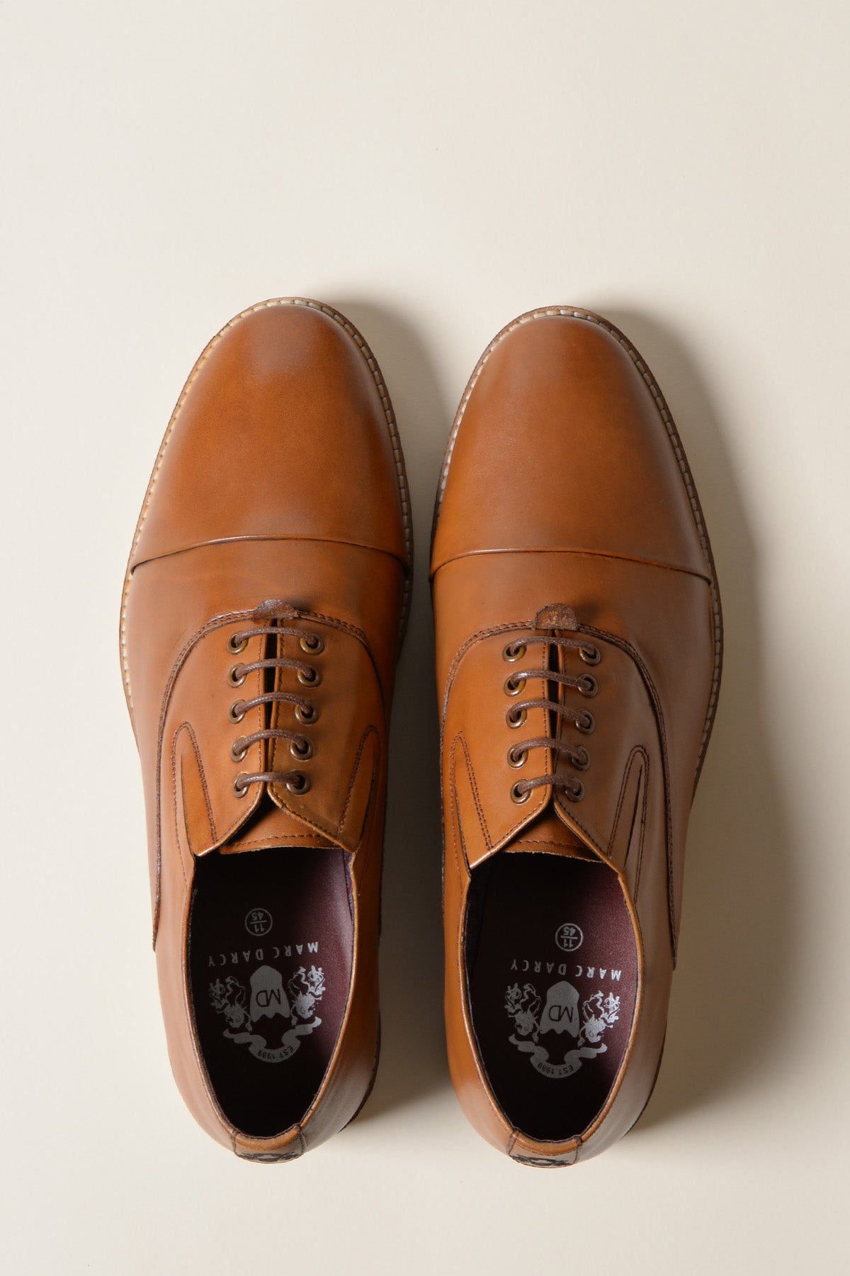 A pair of GAVIN Tan Leather Cap Toe Oxford Shoes by Marc Darcy, viewed from above on a light background. One shoe has tied laces while the other is untied, displaying the brand logo and fine hand-stitched detailing inside.