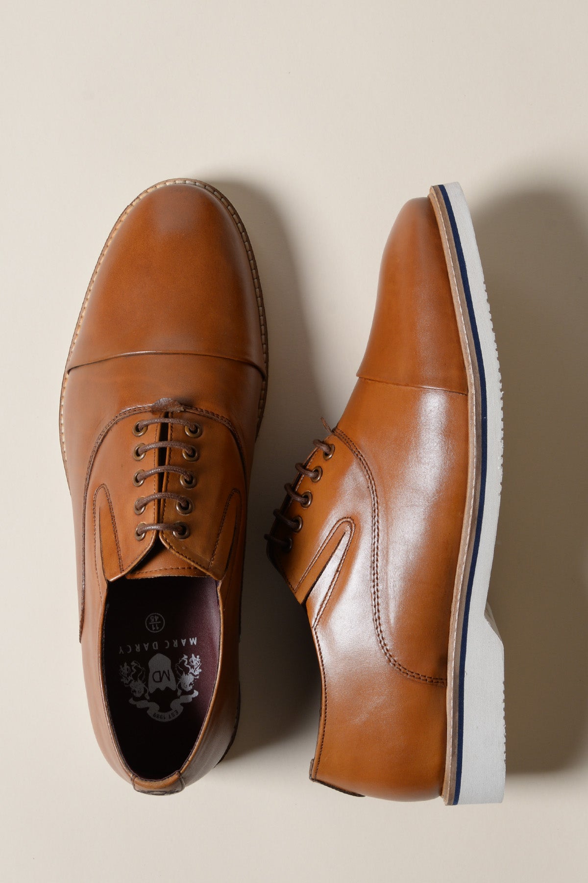 A pair of GAVIN Tan Leather Cap Toe Oxford Shoes by Marc Darcy—one upright showing the side, the other on its side displaying the top and laces—are set against a light beige background.