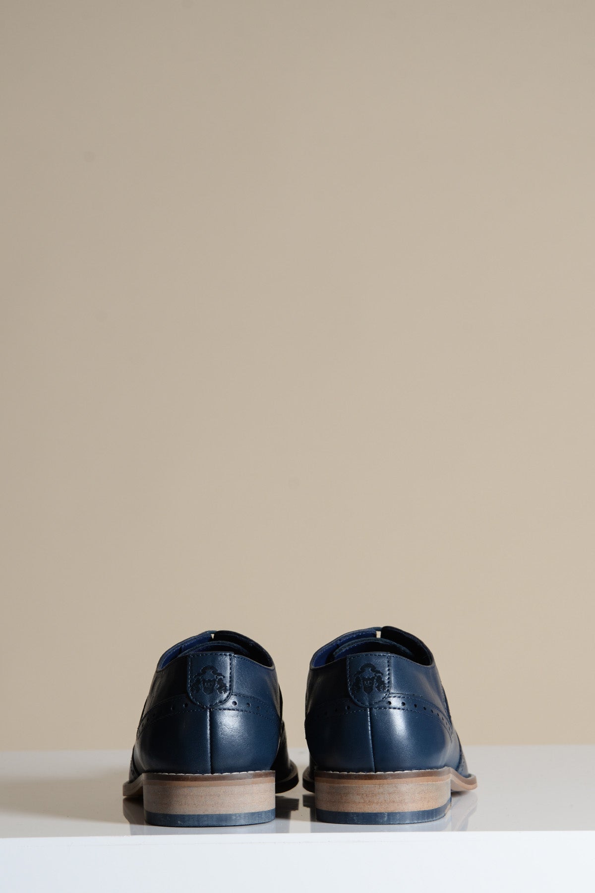The DAWSON Navy Wingtip Oxford Brogue Shoes by Marc Darcy are crafted from full grain leather with brown soles and subtle decorative stitching, shown from the back on a white surface against a beige backdrop.