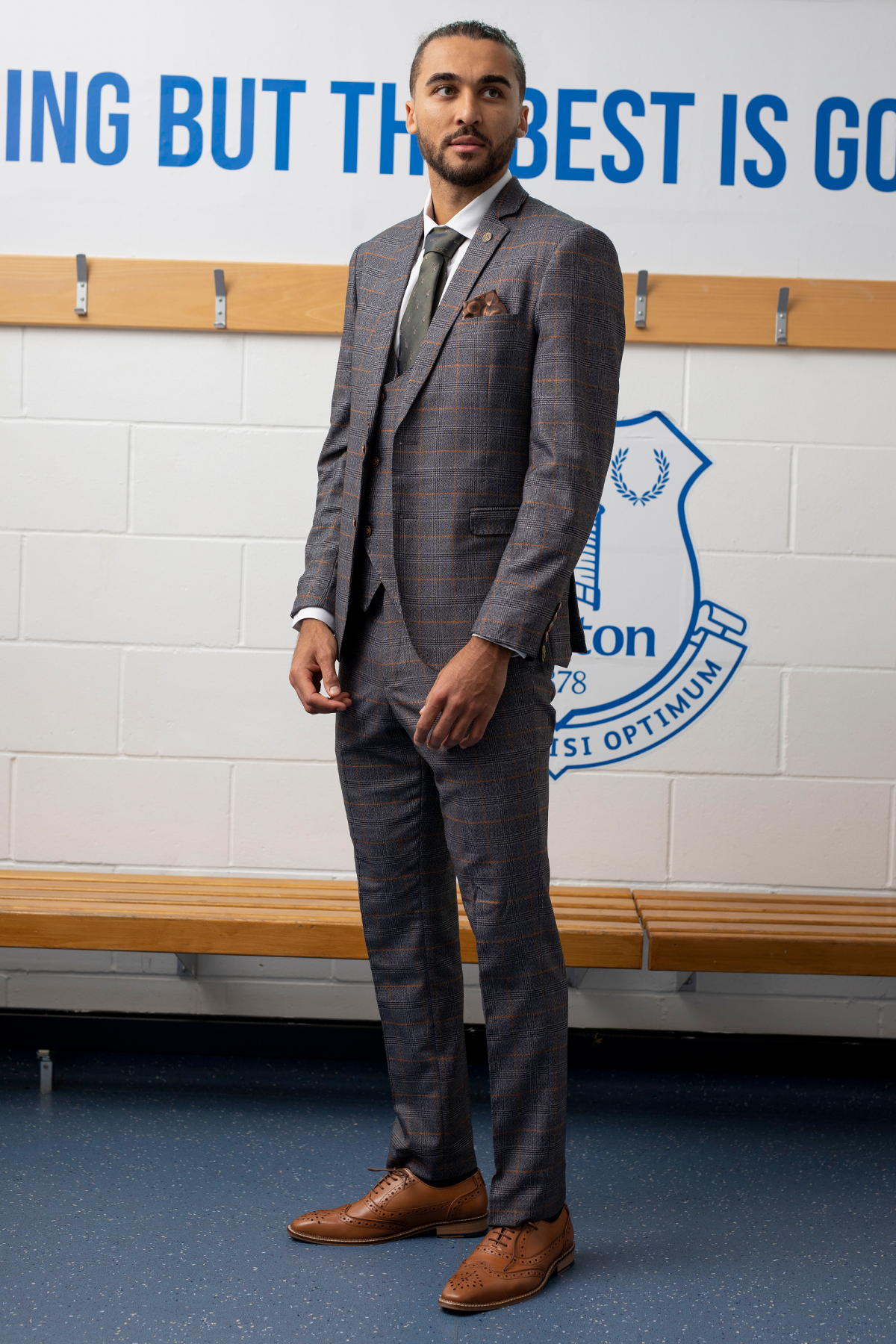 Dominic Calvert-Lewin wears the marcdarcy JENSON Grey Suit from The Everton Collection, standing indoors by a wooden bench with part of a blue and white sports emblem and motivational text on the wall behind him.
