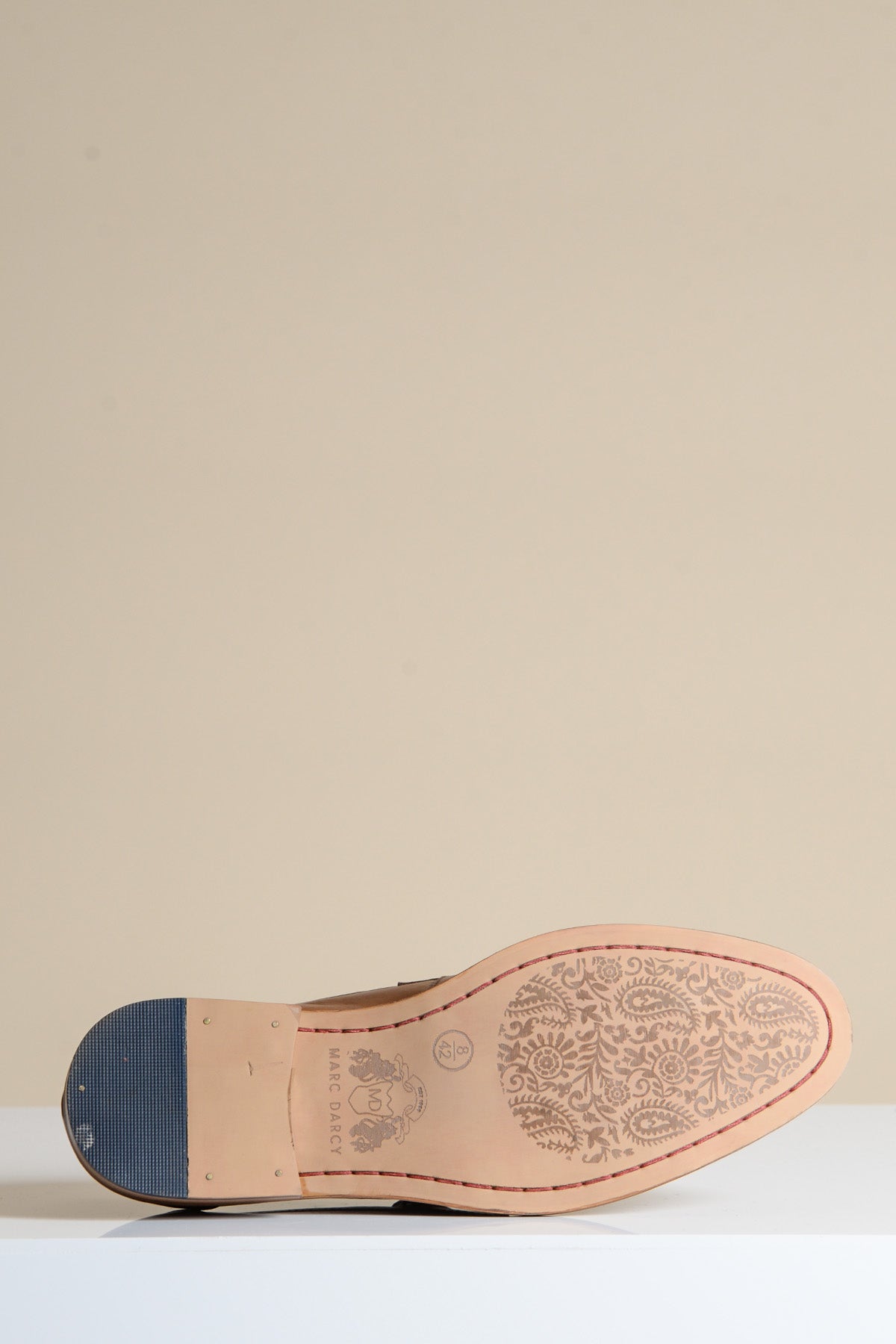 The Marc Darcy JASPER Tan Leather Penny Loafer is displayed sole-up on a white surface, showcasing its full grain leather upper, intricately floral-engraved sole with branding by the heel, and a textured black heel cap. The background is beige.