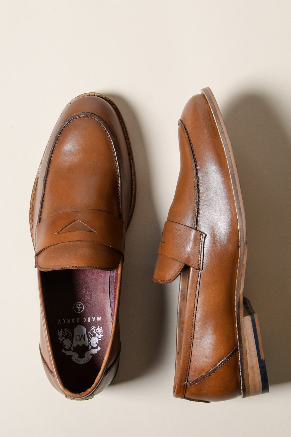 The Marc Darcy JASPER Tan Leather Penny Loafer Shoe, made from full-grain leather with a classic penny slot design, is displayed on a light beige background—one shown from above and one from the side to highlight its stitching and wooden sole.