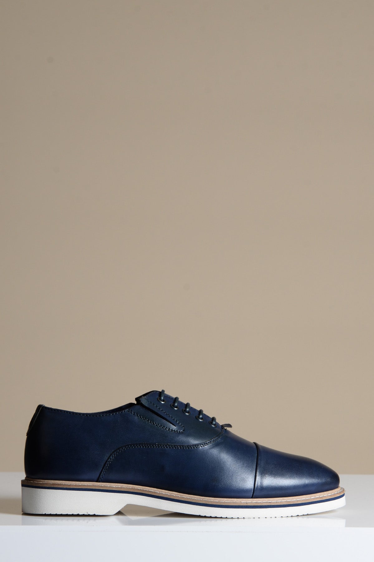 The Marc Darcy GAVIN is a navy blue full grain leather cap toe Oxford shoe with laces and a white sole, shown in profile on a white surface against a plain beige background.