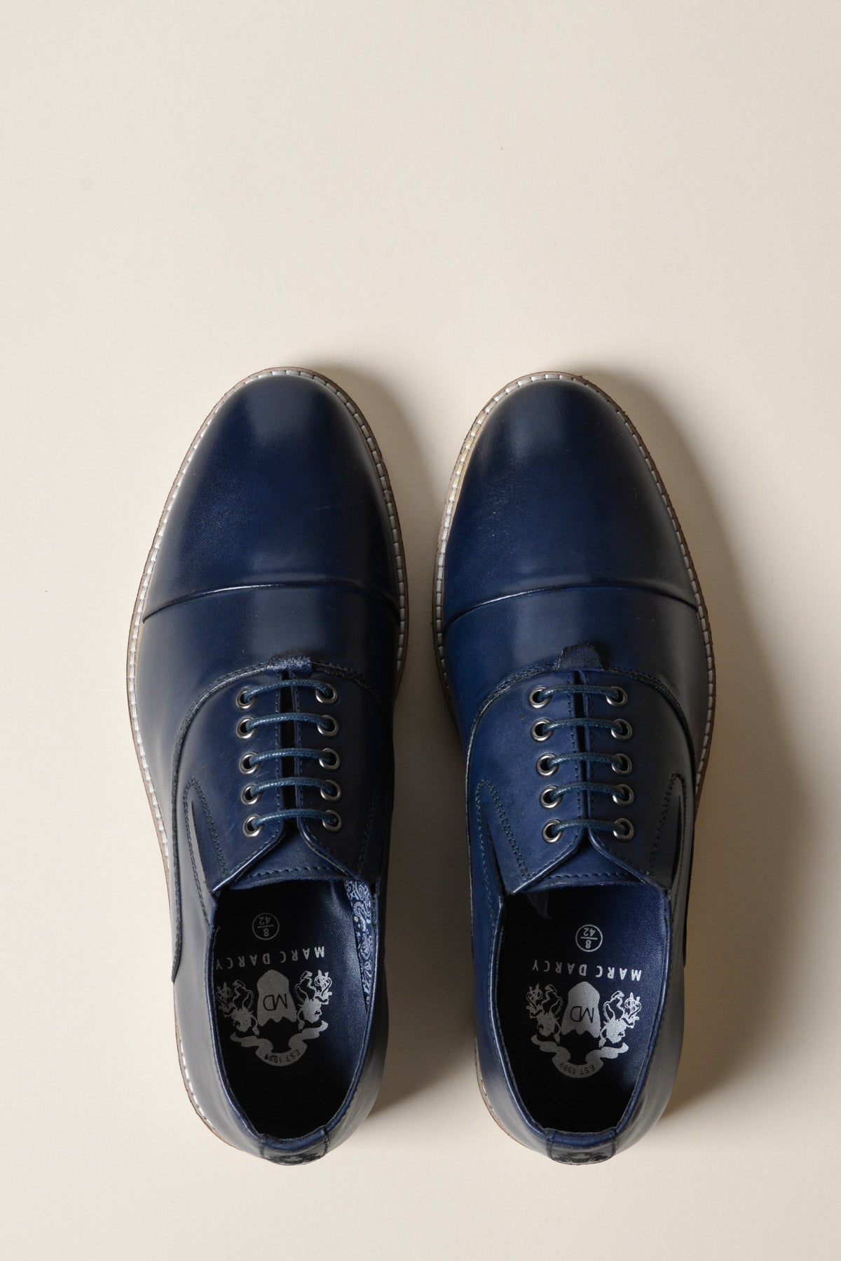A pair of Marc Darcy GAVIN navy blue leather cap toe Oxford shoes with tan soles, shown from above on a light beige background. The left shoe is fully laced; the right has no laces threaded.