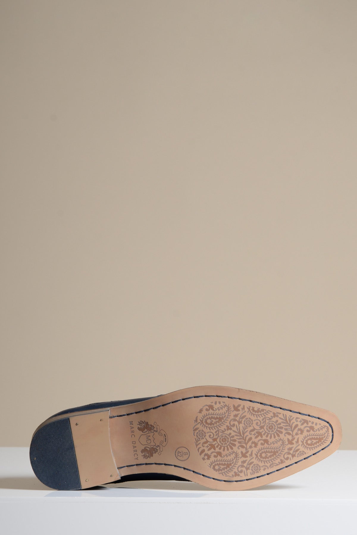 A close-up of the DAWSON Navy Wingtip Oxford Brogue Shoe by Marc Darcy lies on its side, highlighting the tan full grain leather sole with an intricate floral pattern and a black heel against a beige background.