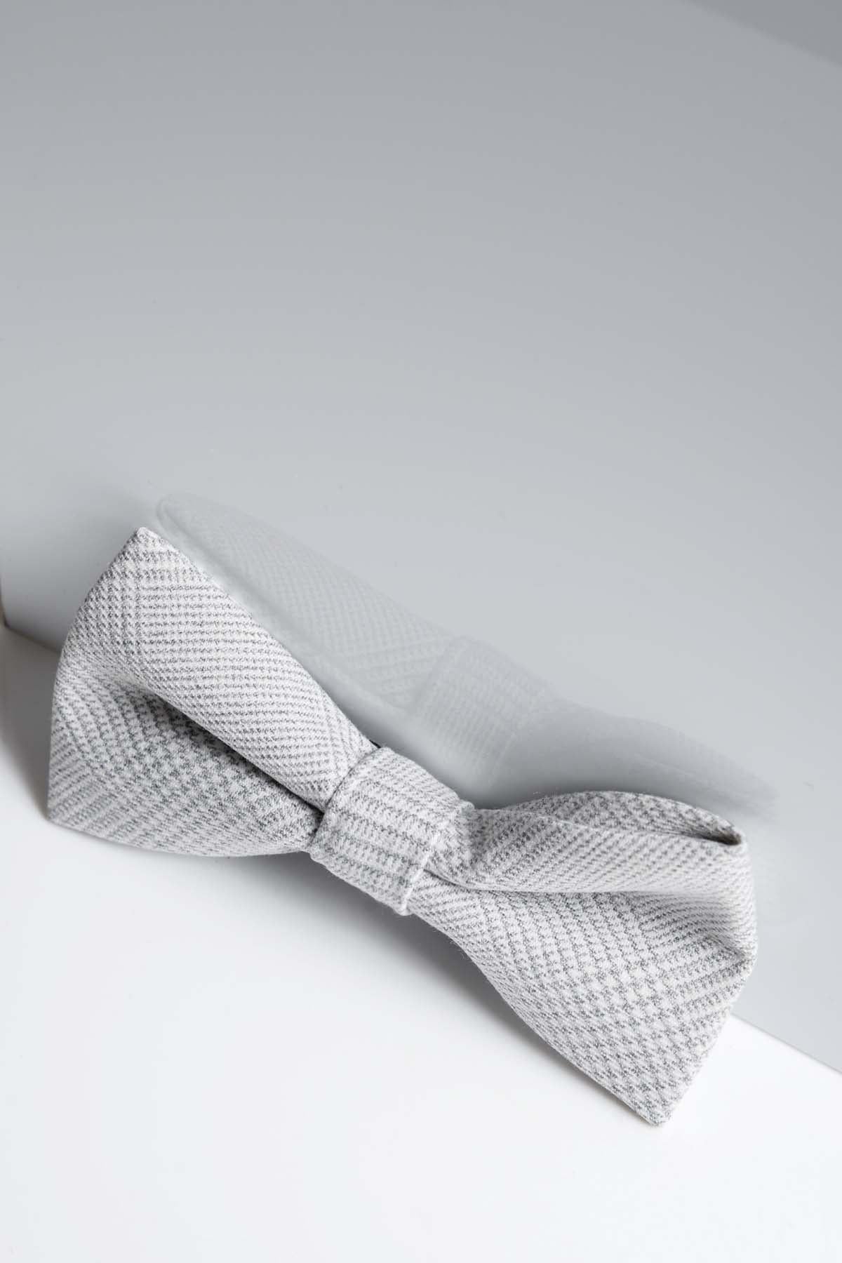 The BROMLEY - Stone Check Bow Tie by Accessory rests on a white surface, its reflection shining above—a stylish light gray option ideal for formalwear and weddings.