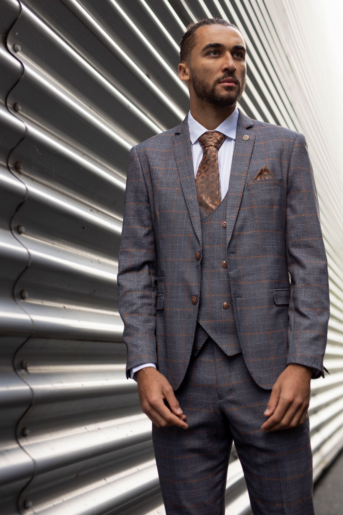 Dominic Calvert-Lewin poses in the marcdarcy JENSON Grey Suit from The Everton Collection, standing confidently by a corrugated metal wall, styled with a brown tie and pocket square.