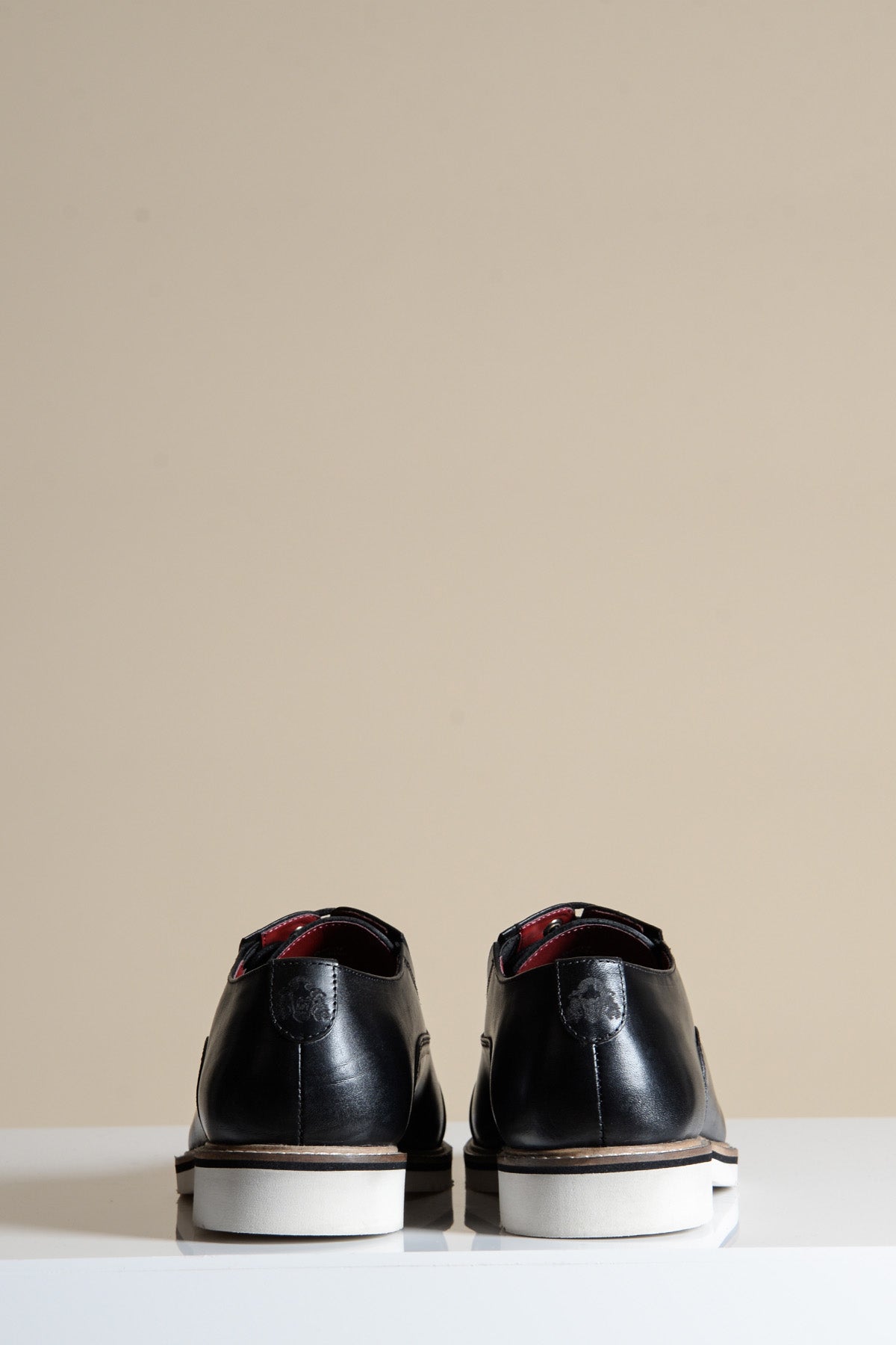 The Marc Darcy GAVIN Black Leather Cap Toe Oxford Shoes are shown from the back, with white soles, placed on a white surface against a plain beige background.