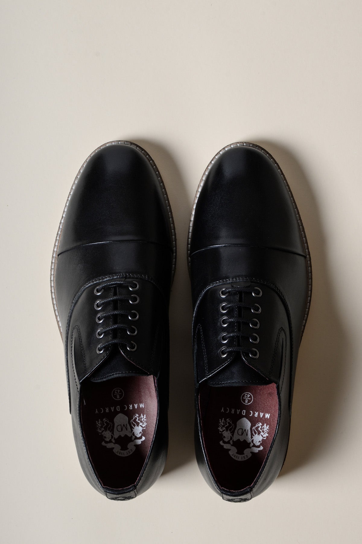 The GAVIN Black Leather Cap Toe Oxford Shoe by Marc Darcy features classic style, white stitching, and silver eyelets, shown from above on a light cream background—formal footwear with impeccable detail.
