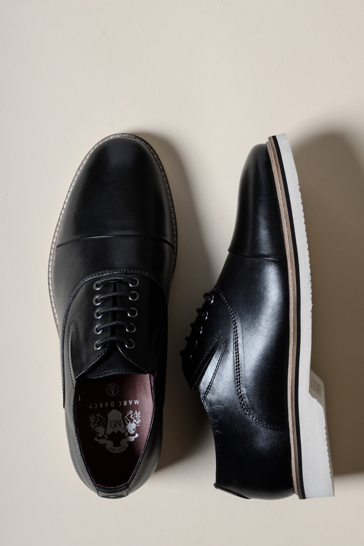 The GAVIN Black Leather Cap Toe Oxford Shoes by Marc Darcy are displayed on a light beige background—one shoe upright to show the laces, the other on its side. These full grain leather lace-ups feature contrasting white soles.