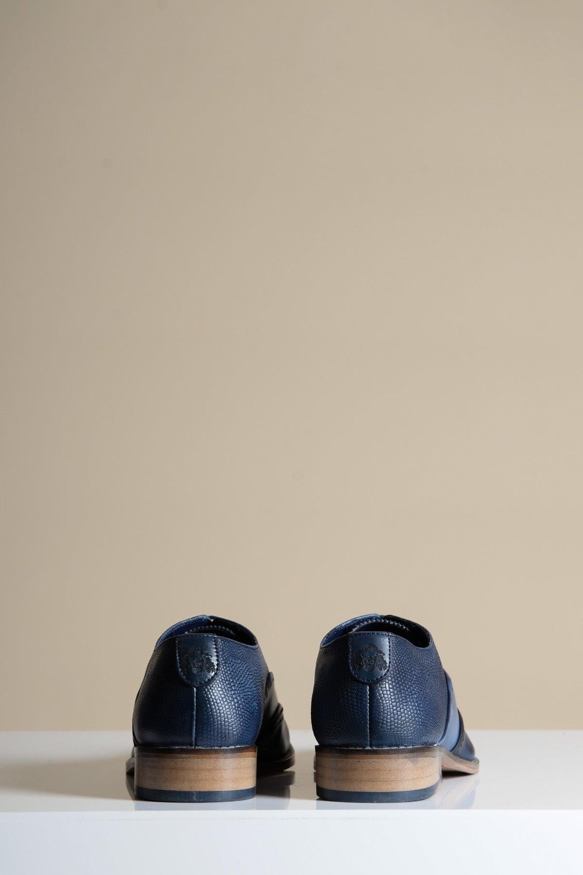 The Marc Darcy RYAN Navy Blue Leather Wingtip Brogue shoes, featuring light brown soles, are shown from the back on a white surface with a beige background.