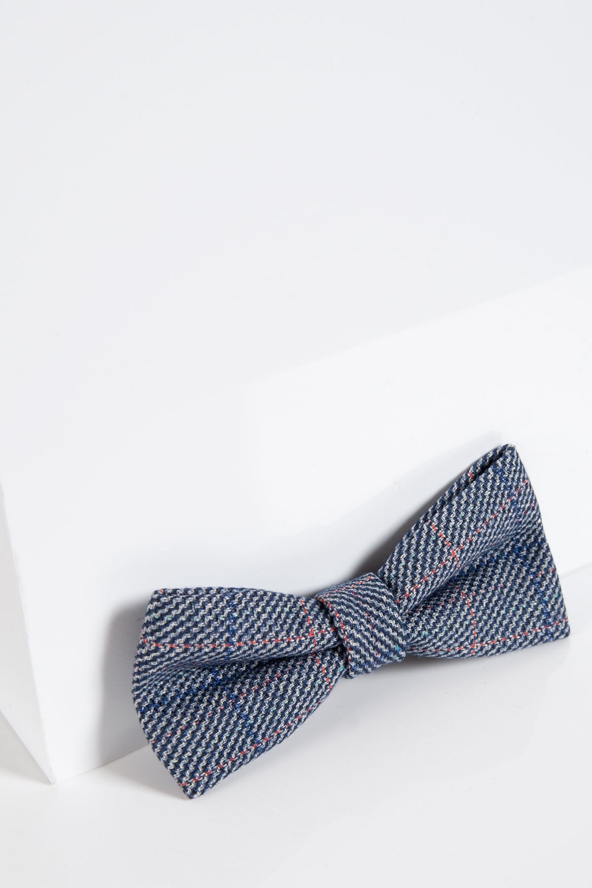 The Accessory HILTON Blue Check Tweed Bow Tie, in a classic butterfly shape with red and white accents, is displayed on a white surface by the corner of a white wall.