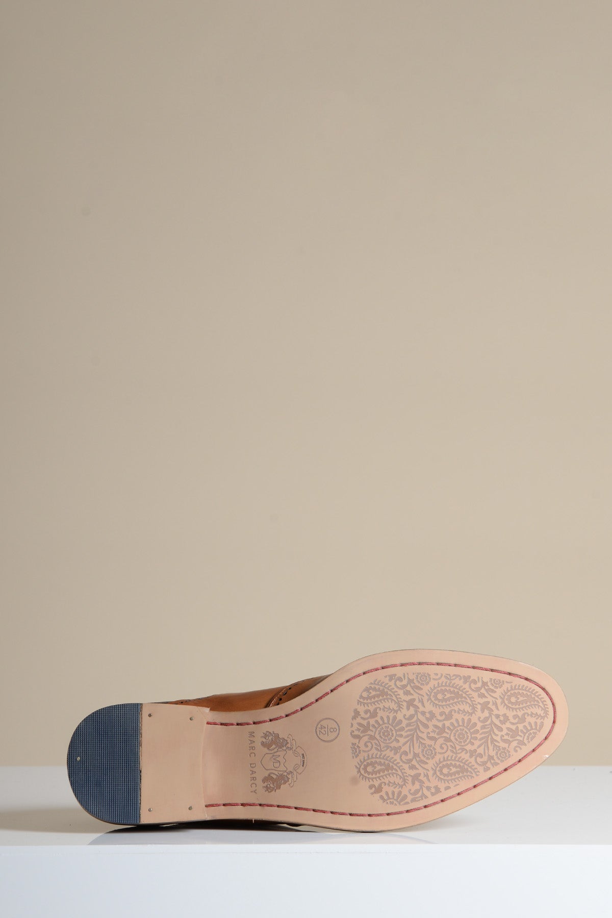 The Marc Darcy RYAN - Tan Brown Leather Wingtip Brogue Shoe lies on its side, showing its floral-patterned sole, blue rubber heel, and hand-stitched details against a plain beige background on a white surface.