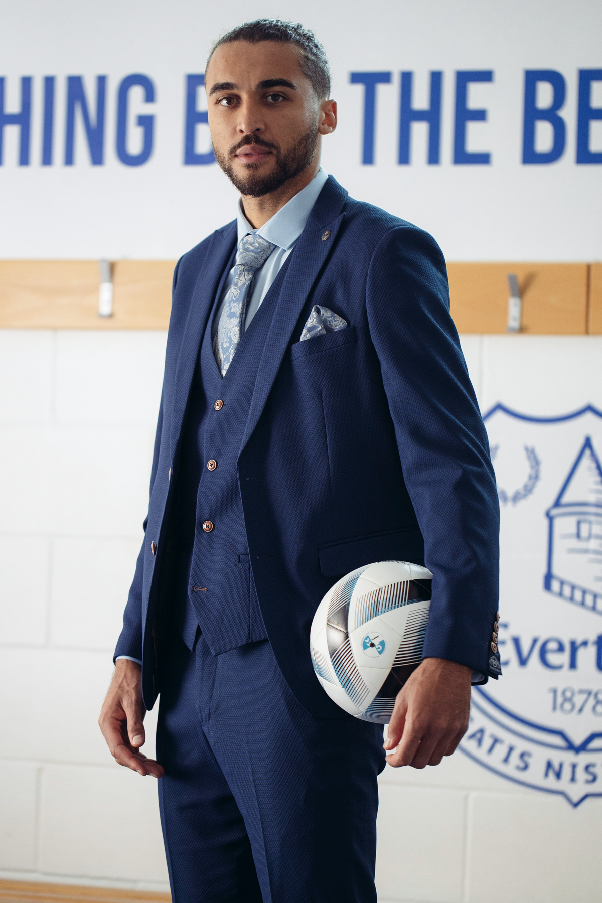 Dominic Calvert-Lewin wears the marcdarcy MAX Royal Blue Suit from The Everton Collection, paired with a patterned tie and camouflage pocket square, holding a soccer ball before a wall featuring motivational text and an Everton team logo.