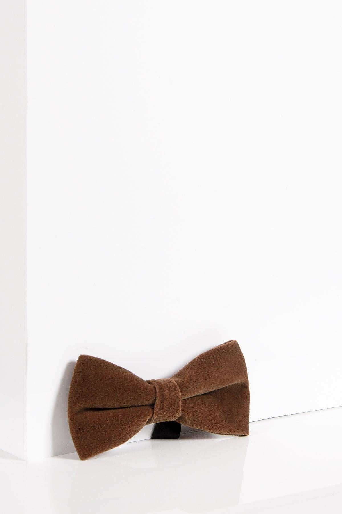 The Accessory VELVET - Dark Tan Velvet Bow Tie, featuring an adjustable strap, is displayed on a white background near the corner where two walls meet.