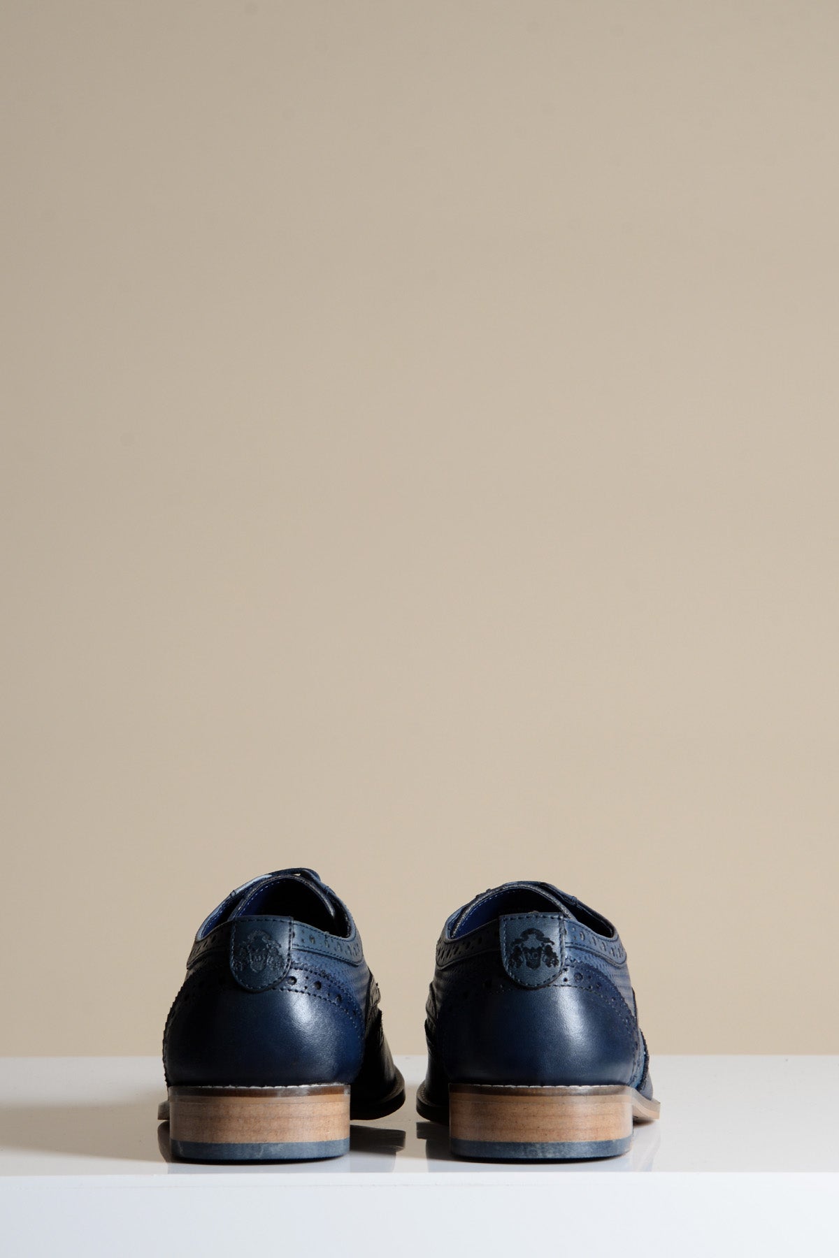 The marcdarcy BRANDON is a navy blue leather wingtip brogue shoe featuring a hand-crafted upper and light brown soles, displayed from the back on a white surface with a plain beige background.