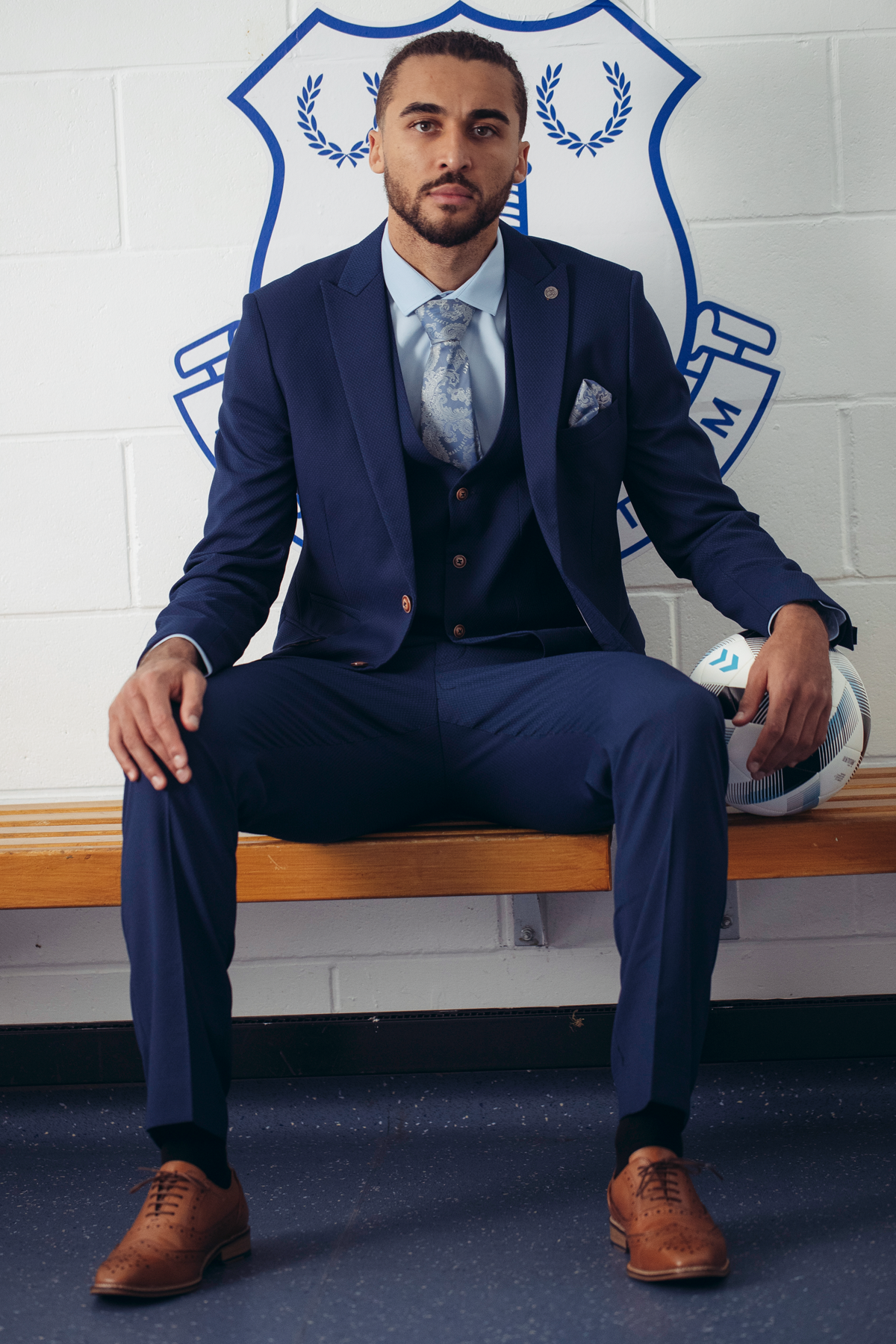 Dominic Calvert-Lewin wears The Everton Collection | MAX Royal Blue Suit by marcdarcy, sitting on a bench with a soccer ball, in front of a white wall with an Everton crest. He pairs the suit with brown dress shoes and looks confidently at the camera.