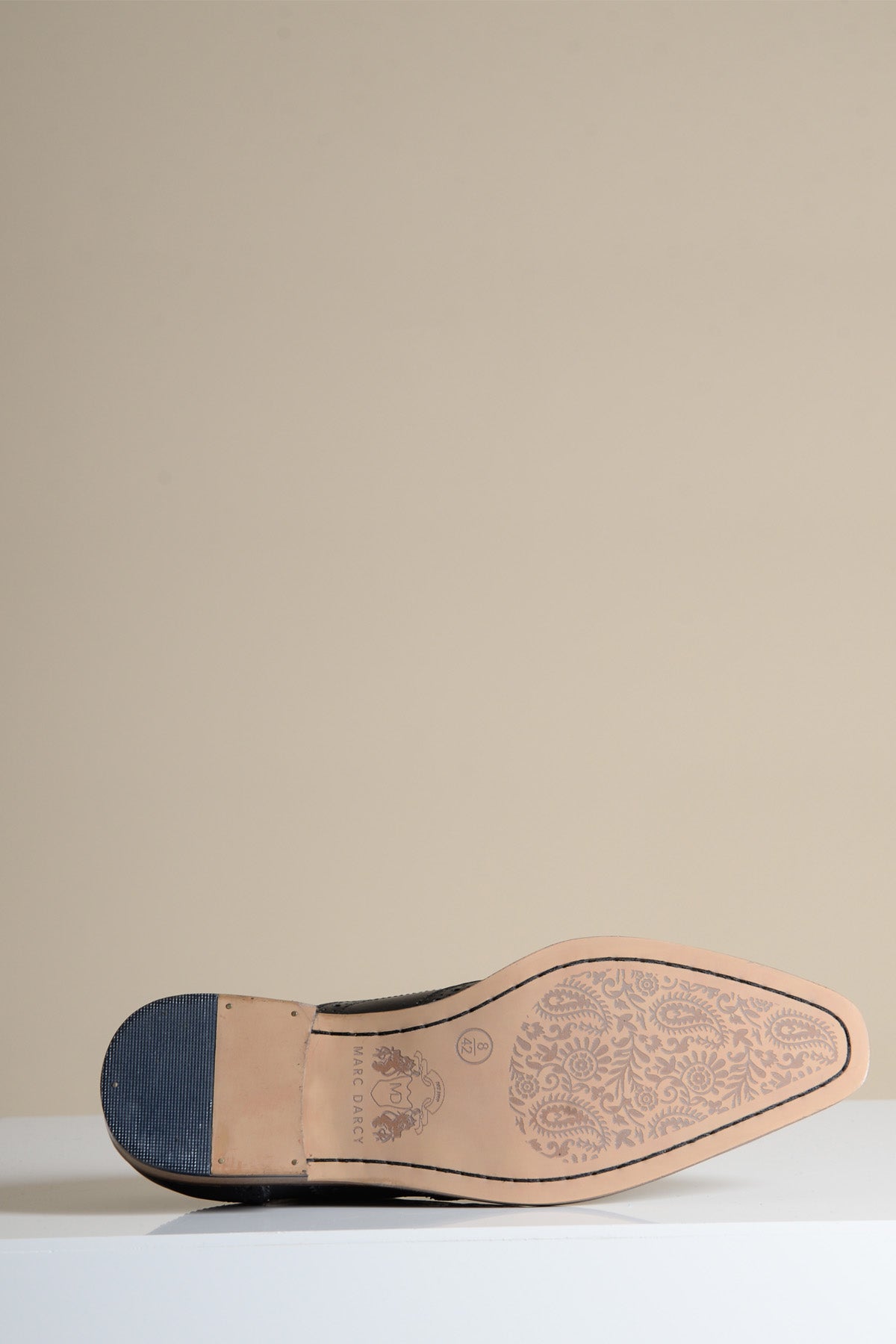 The MARCO Black Grey Leather Wingtip Oxford Shoe by Marc Darcy is shown upside down, revealing its tan sole adorned with an intricate floral pattern, set against a plain beige background.