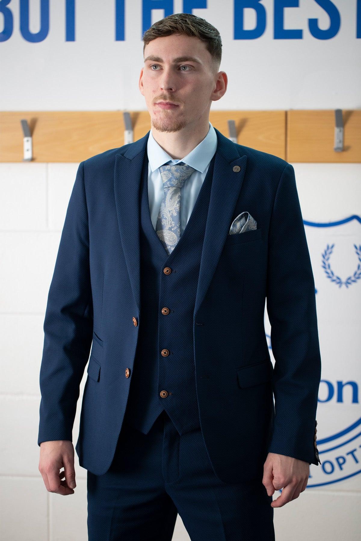 A man wears the marcdarcy Everton Collection | James Garner in the MAX Royal Blue Suit with a patterned tie and pocket square, standing indoors before a wall with coat hooks and blue text.